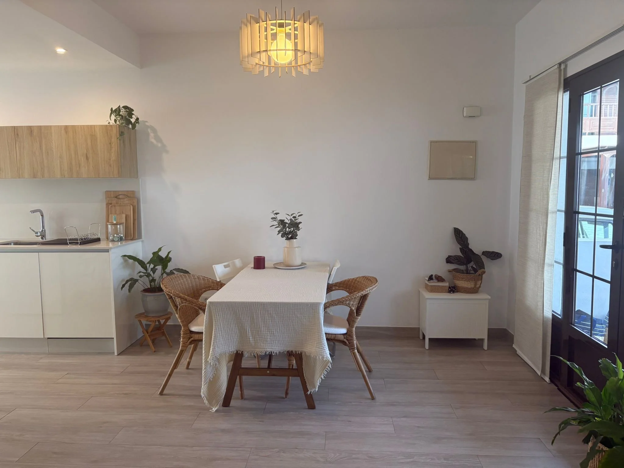 Modern dining area with a wooden table and wicker chairs, white tablecloth, potted plants, and a minimalistic kitchen in the background. Wooden cabinets, a sink, and cutting boards are visible. Natural light enters through a glass door alongside indo