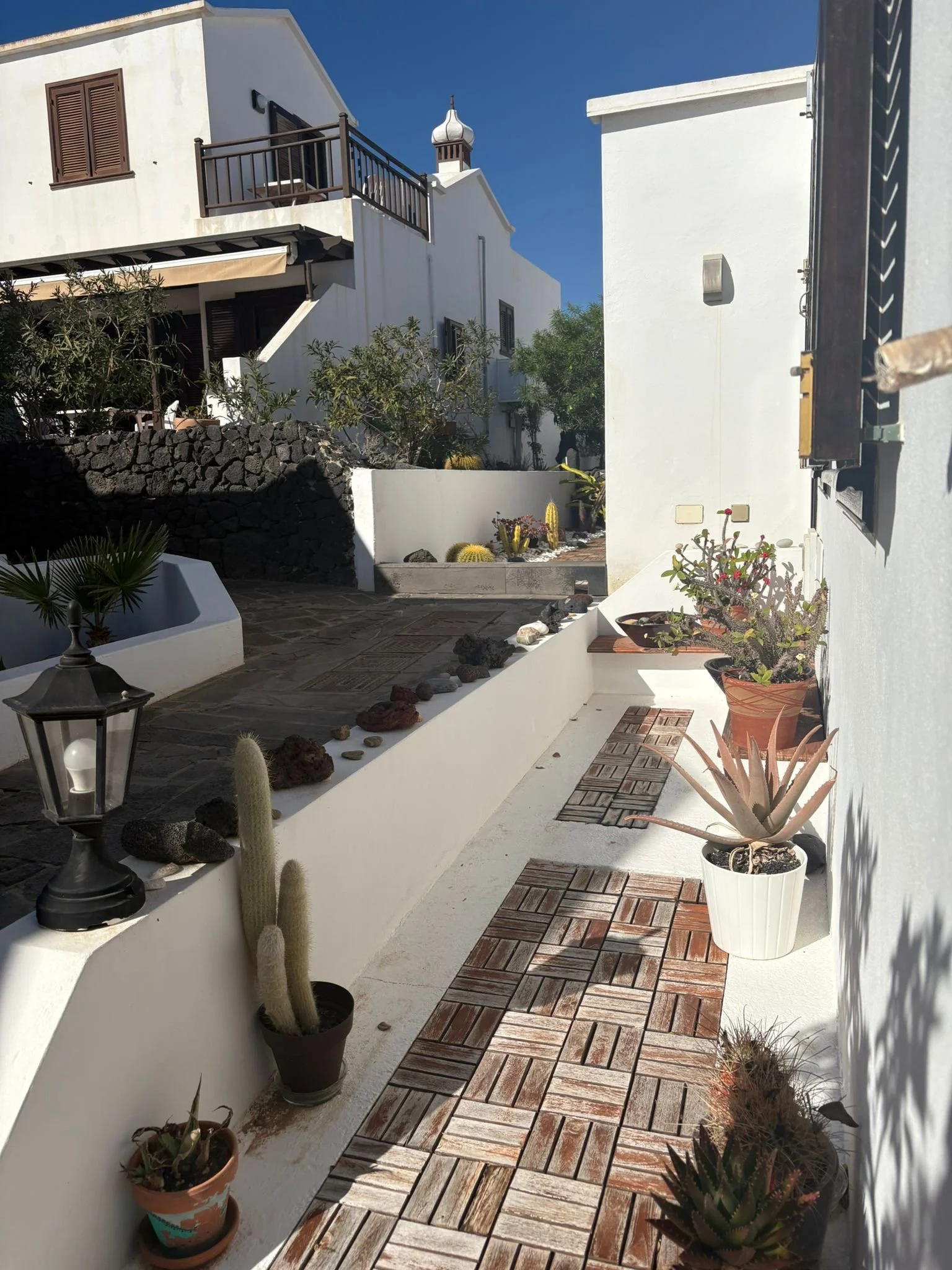 White house with a wooden balcony and brown shutters, surrounded by a garden with various potted plants and cacti. A paved pathway with wooden tiles and small decorative stones runs alongside a white wall. A lamppost and a tower feature are visible a