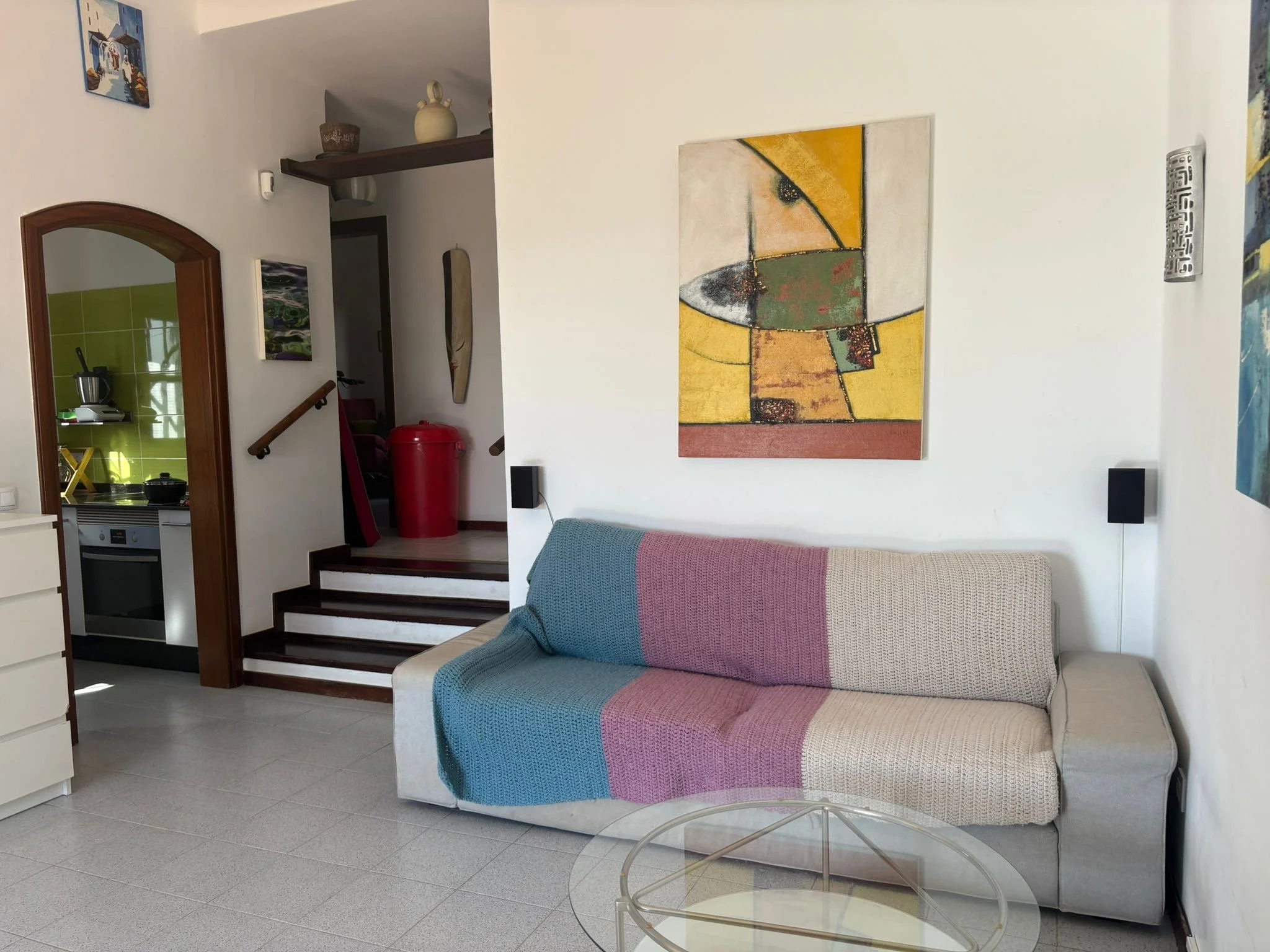 Modern living room with abstract art, a beige and purple knitted throw on a sofa, a glass coffee table, and a view into the kitchen with a green backsplash and oven. A staircase with a red trash bin is visible in the background.