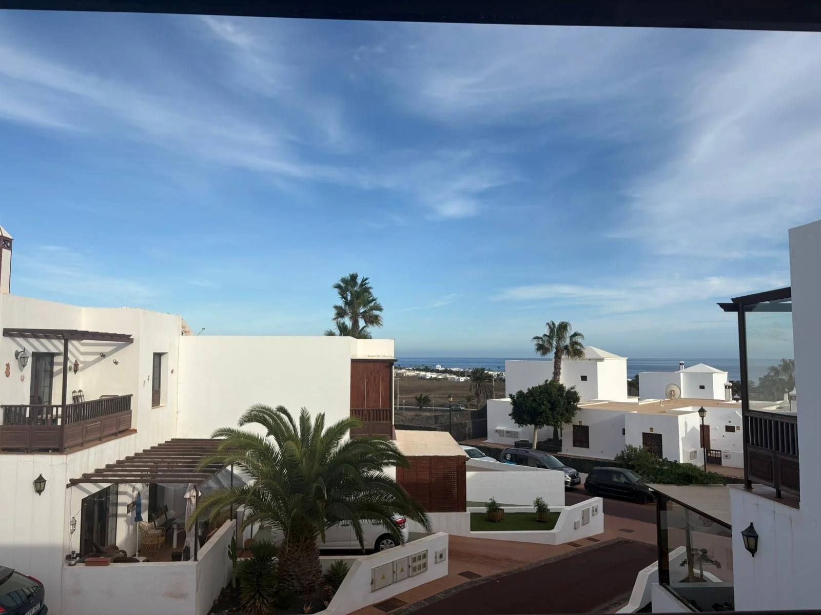 View of a coastal residential area with white buildings, palm trees, and the ocean in the background under a blue sky with scattered clouds.