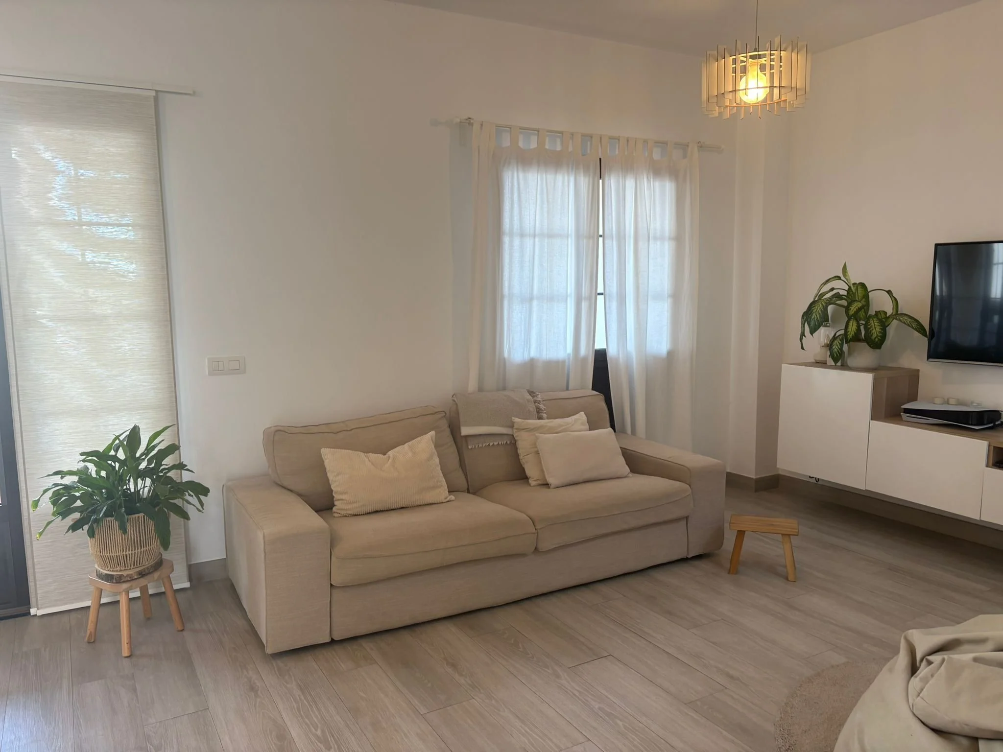 Modern living room with beige sofa, white curtains, potted plants, TV on wall, and wooden flooring.