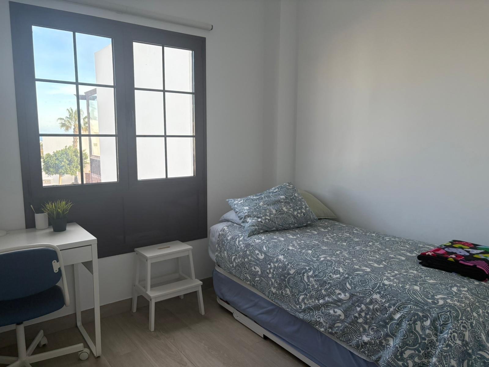 Small bedroom with a single bed, desk, and chair, featuring a large window showcasing an outdoor view with trees.