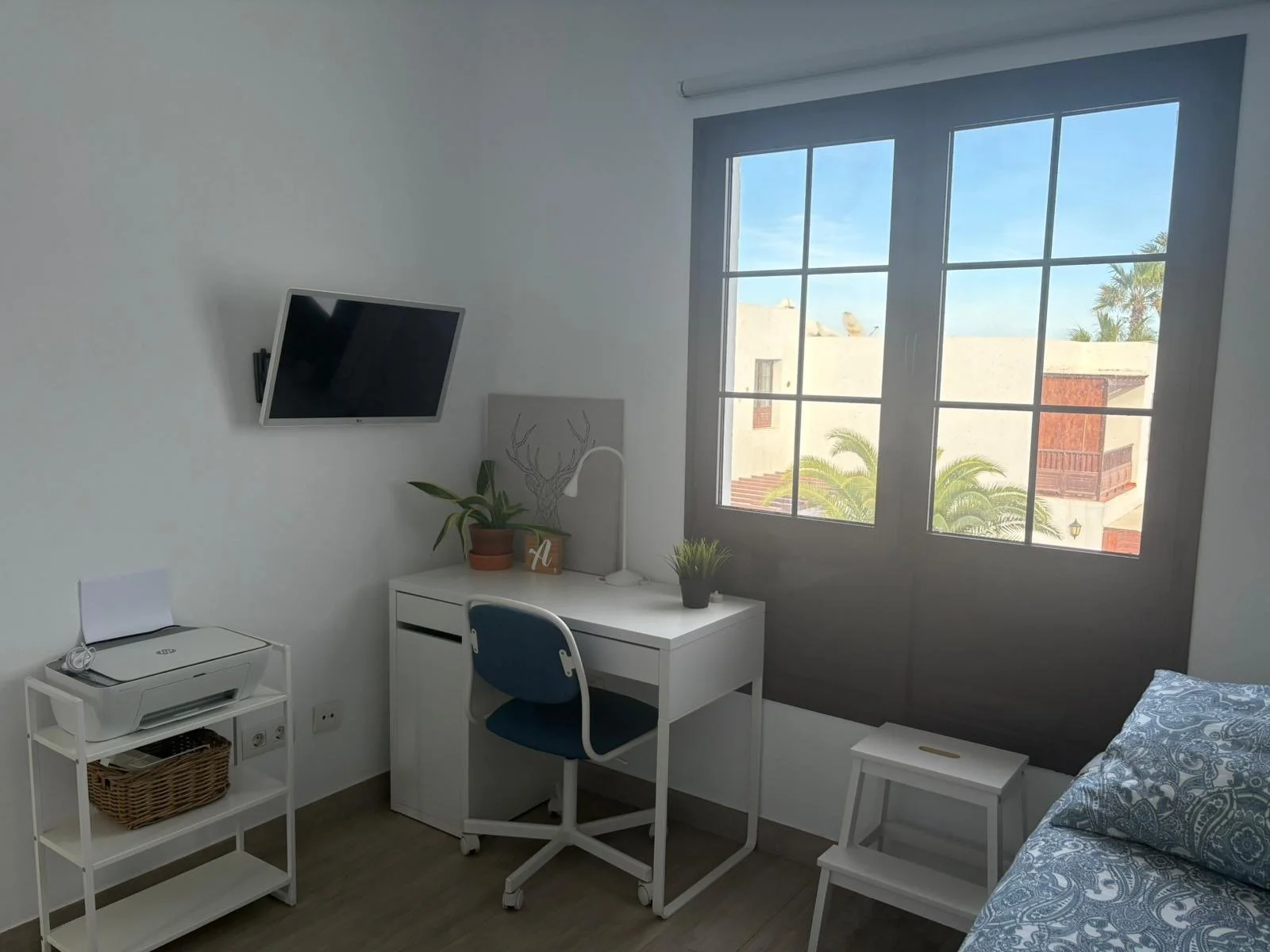 A modern, minimalist bedroom with a desk and a large window. The room has a small white desk with a potted plant, chair, and a wall-mounted TV. There's a printer on a shelf beside the desk. The window offers a view of a building and trees outside. A 