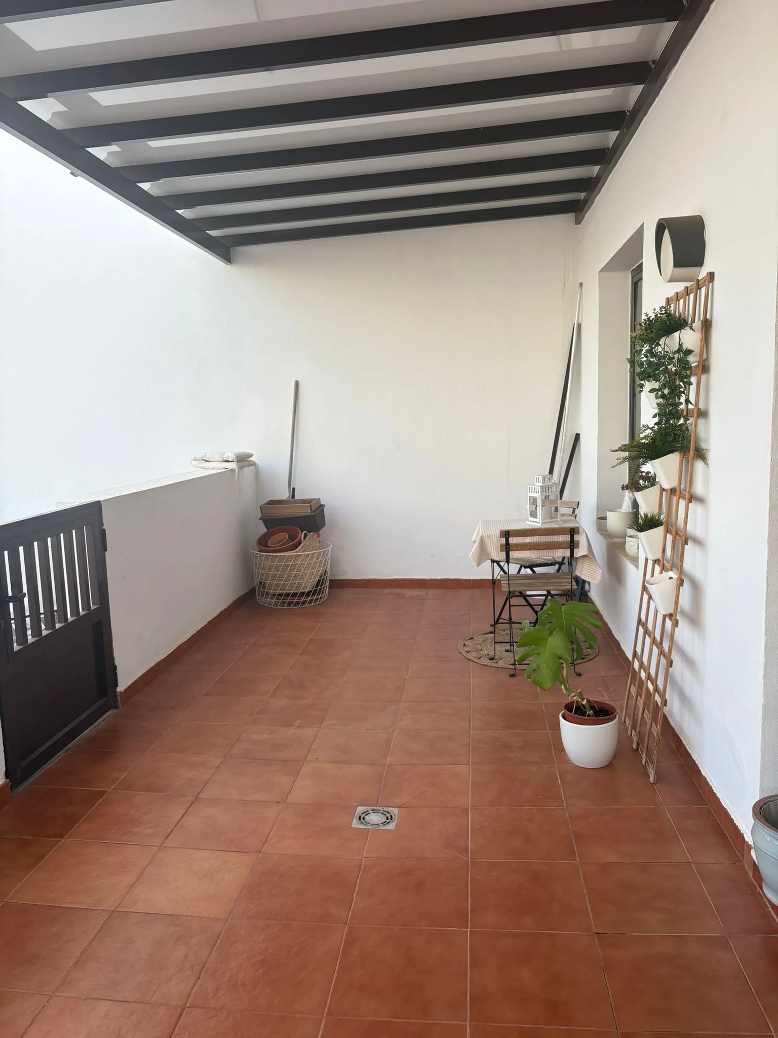 Covered patio with terracotta tiles, small table with a chair, potted plants, trellis with plants, a basket with pots, and broom against white walls.