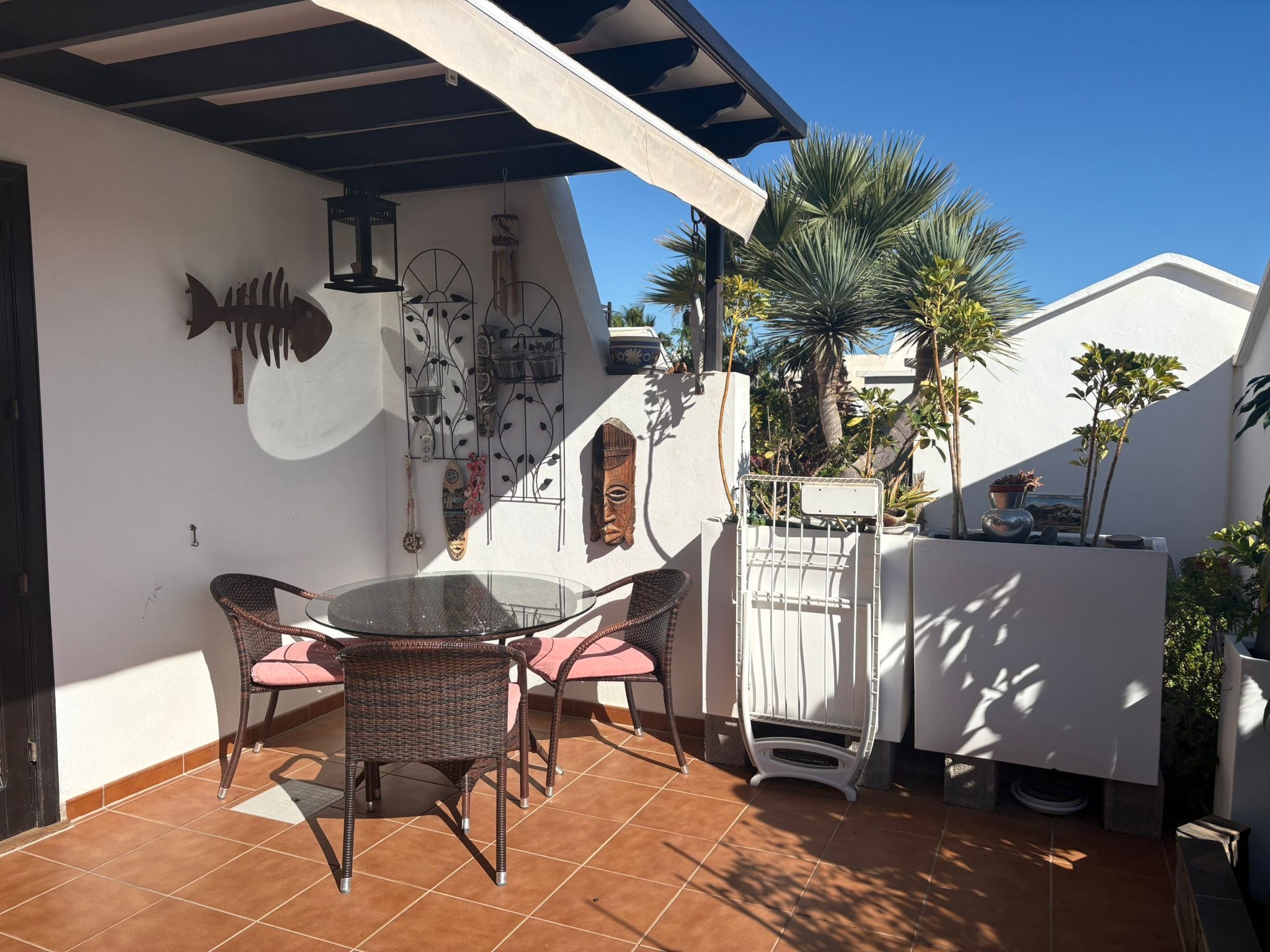 Outdoor patio with wicker chairs and a glass table, decorative wall art, and plants. Terracotta floor tiles and white walls. Clear blue sky background.