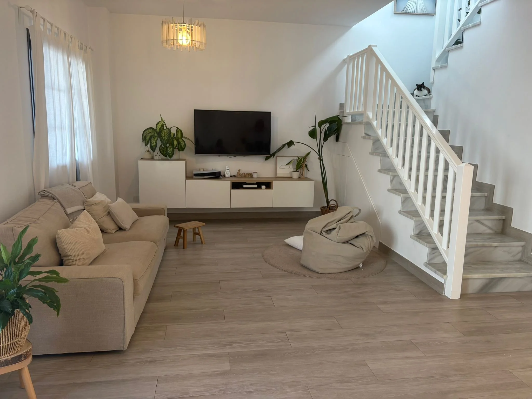 Modern living room with beige couch, TV on wall, white media console, indoor plants, stairs with cat, hanging light fixture, wood flooring, and gray bean bag chair.