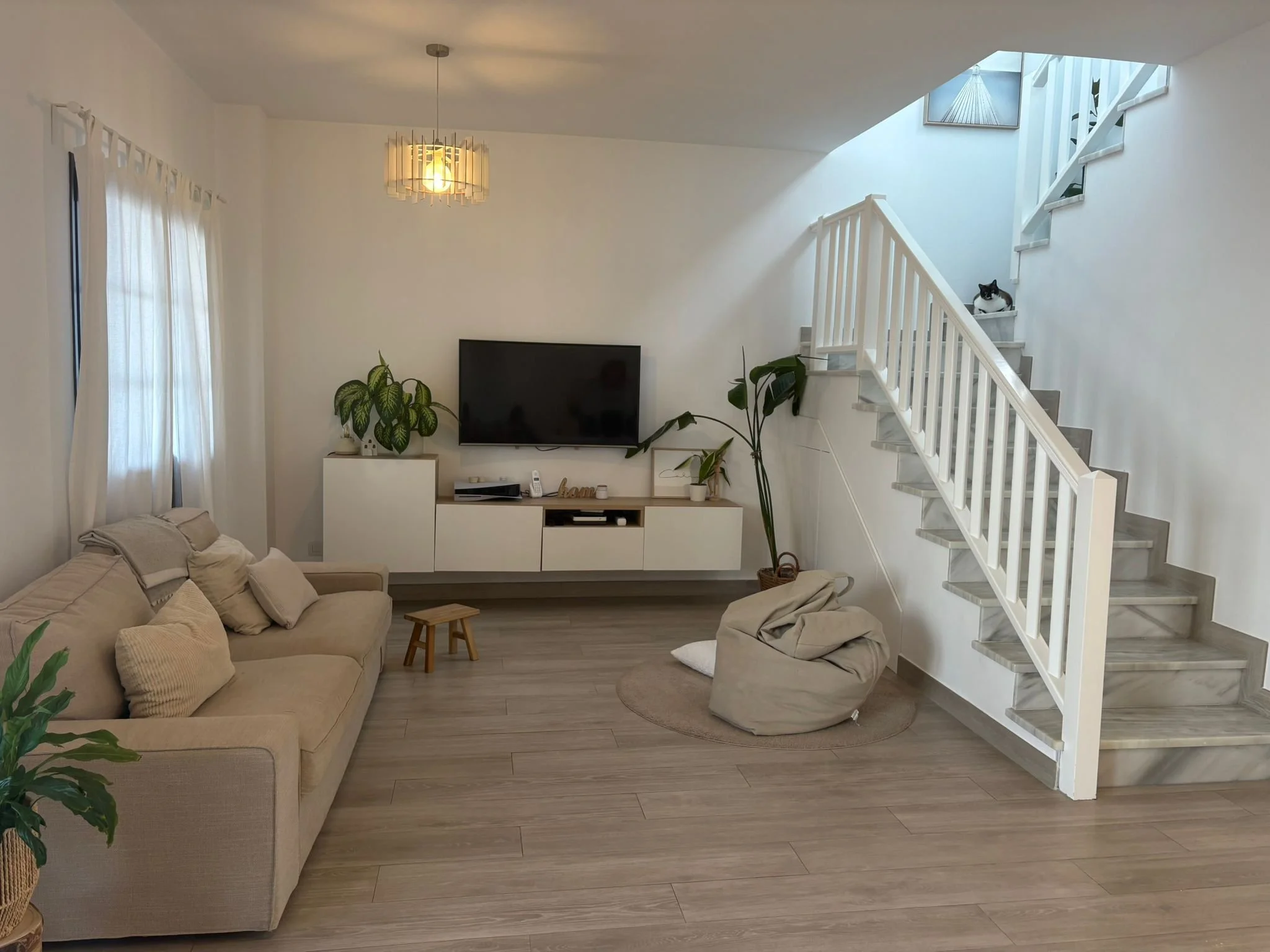 Modern living room with a beige sofa, decorative pillows, and a small wooden stool. A television is mounted on the wall above a white entertainment unit, flanked by green potted plants. A staircase with white railings is visible, with a cat resting o