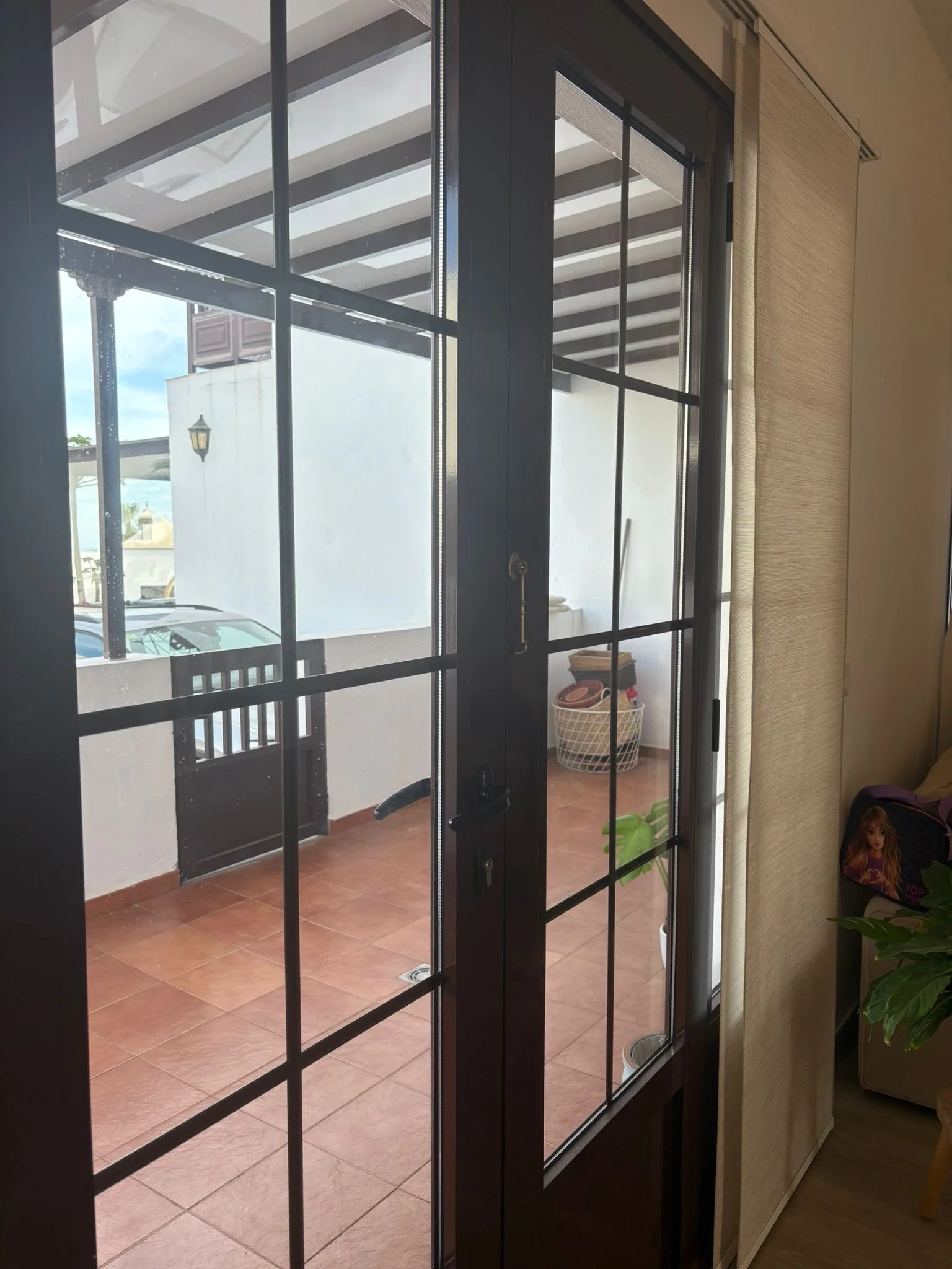 Glass door overlooking a tiled outdoor patio with a small basket containing items, a visible portion of a car, and a decorative wall light. Indoor plants are partially visible inside.