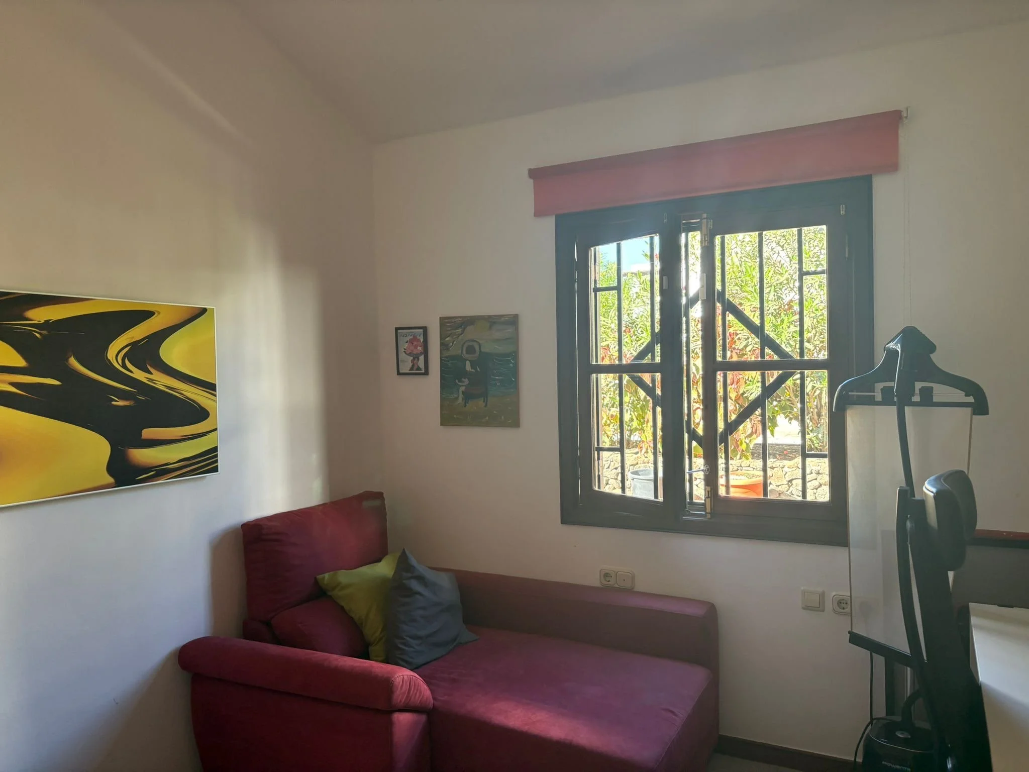 Room with a red sofa, abstract yellow and black painting, two smaller wall artworks, window with bars, and clothing valet stand.