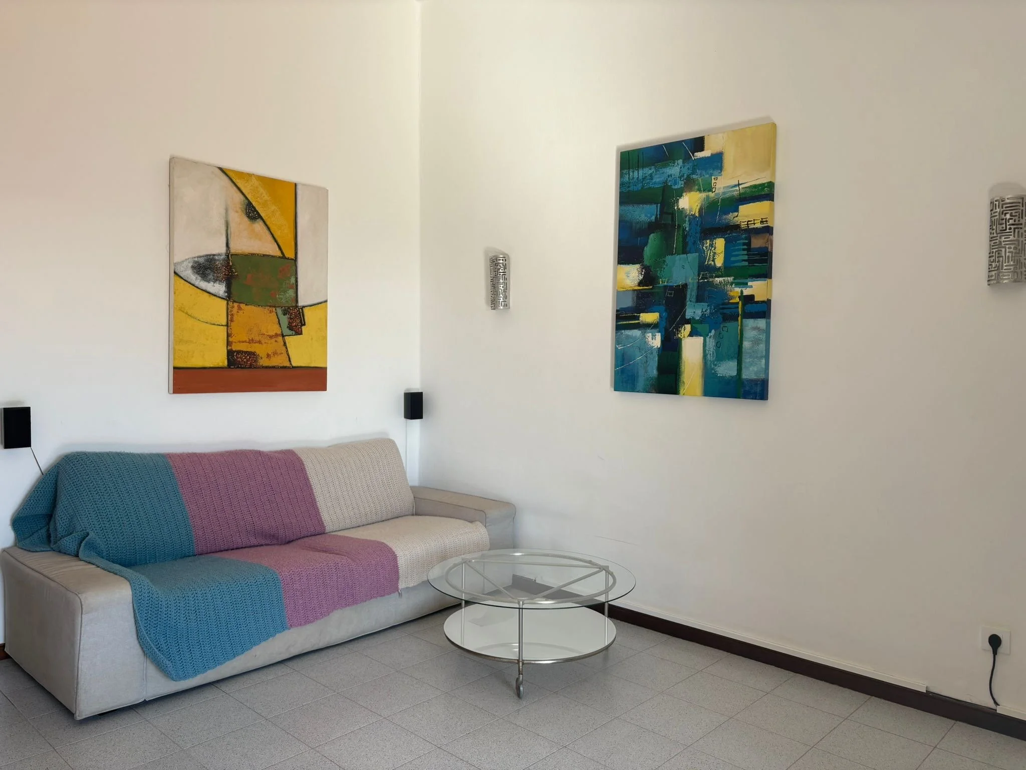 Living room with abstract paintings on the wall, a beige sofa covered with blue and purple blankets, a round glass coffee table, and wall-mounted lights.