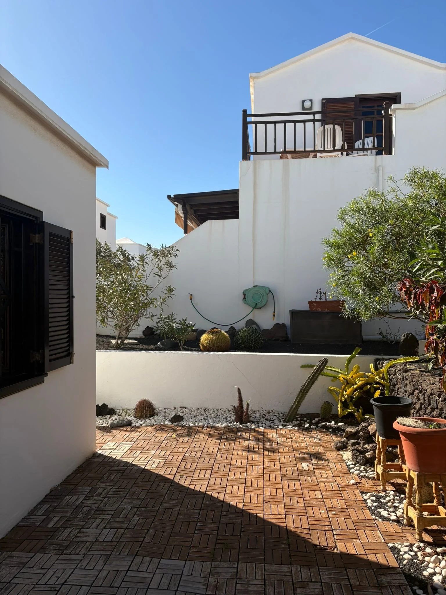 Outdoor patio area with a wooden deck and white walls, featuring several potted plants including cacti and succulents. A balcony with chairs is visible on the second floor. Clear blue sky overhead.
