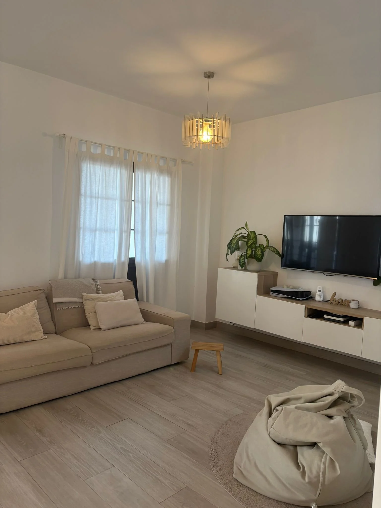 Modern living room with beige sofa, pillows, large window with white curtains, ceiling light, wall-mounted TV, wooden floor, bean bag, small wooden stool, and a shelf with plants and decor items.