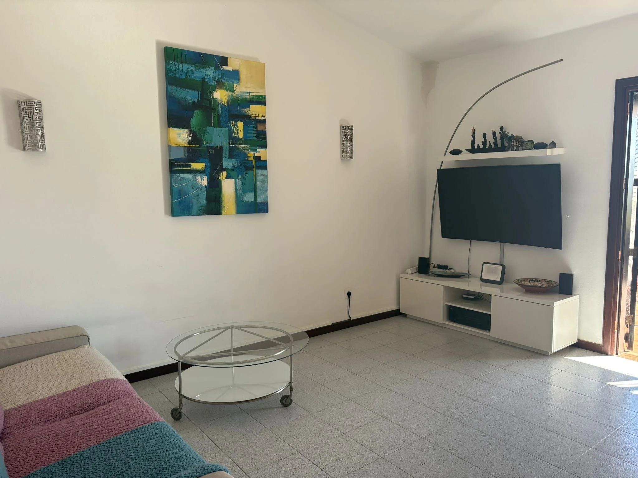 A modern living room with a colorful abstract painting on the wall, a white entertainment unit with a TV, and a decorative shelf. There is a glass coffee table and a cozy sofa with a multicolored throw blanket.
