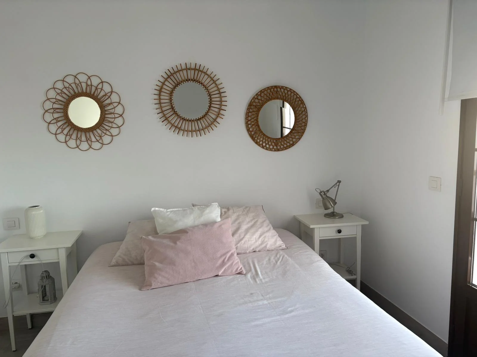 Minimalistic bedroom with a bed, pink and white bedding, decorative mirrors on the wall, and two white nightstands with a vase and lamp.