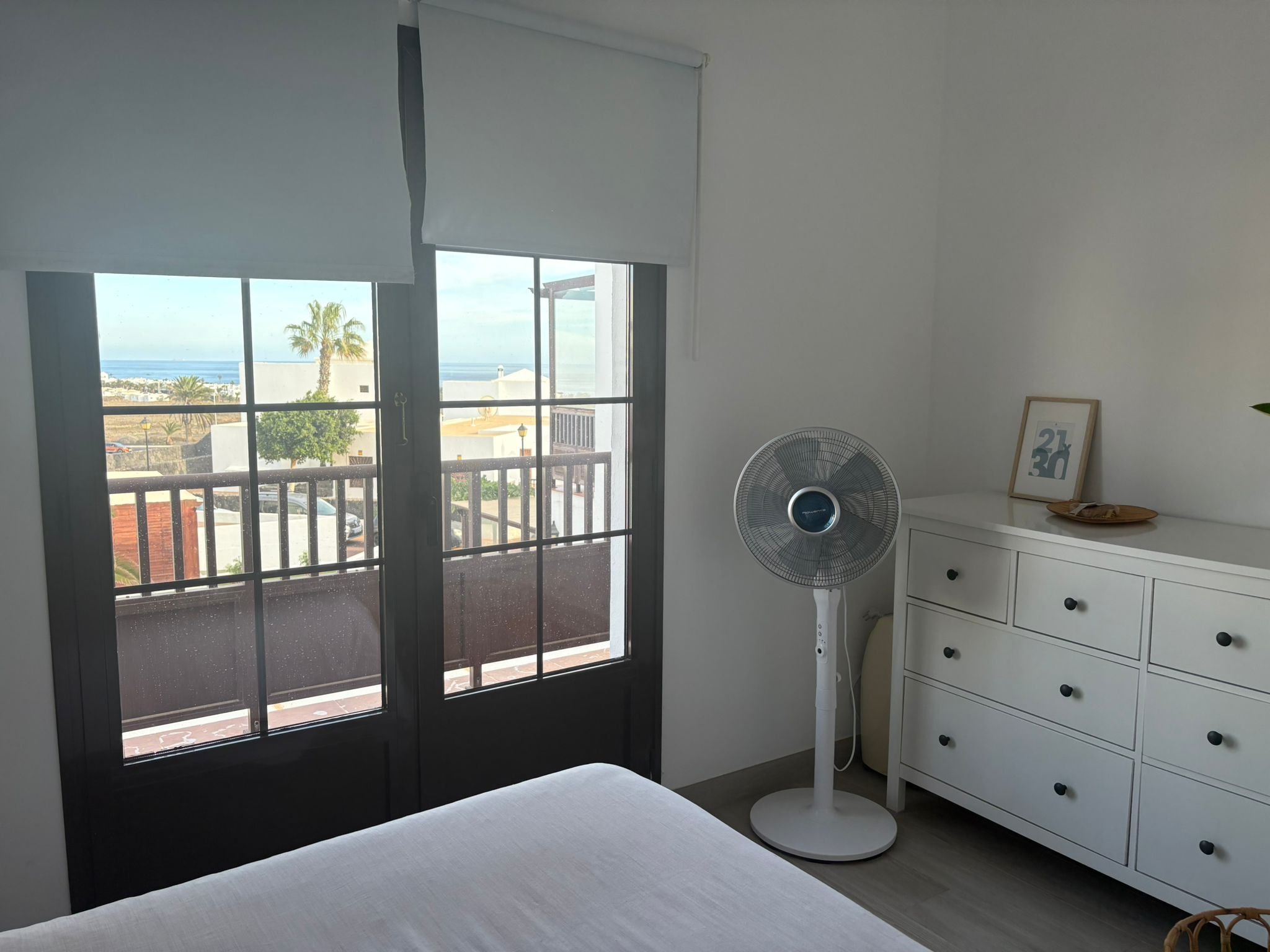 Bright bedroom with a large window, light-colored bed, white dresser, standing fan, and framed artwork, showcasing an outdoor view with palm trees and the sea.