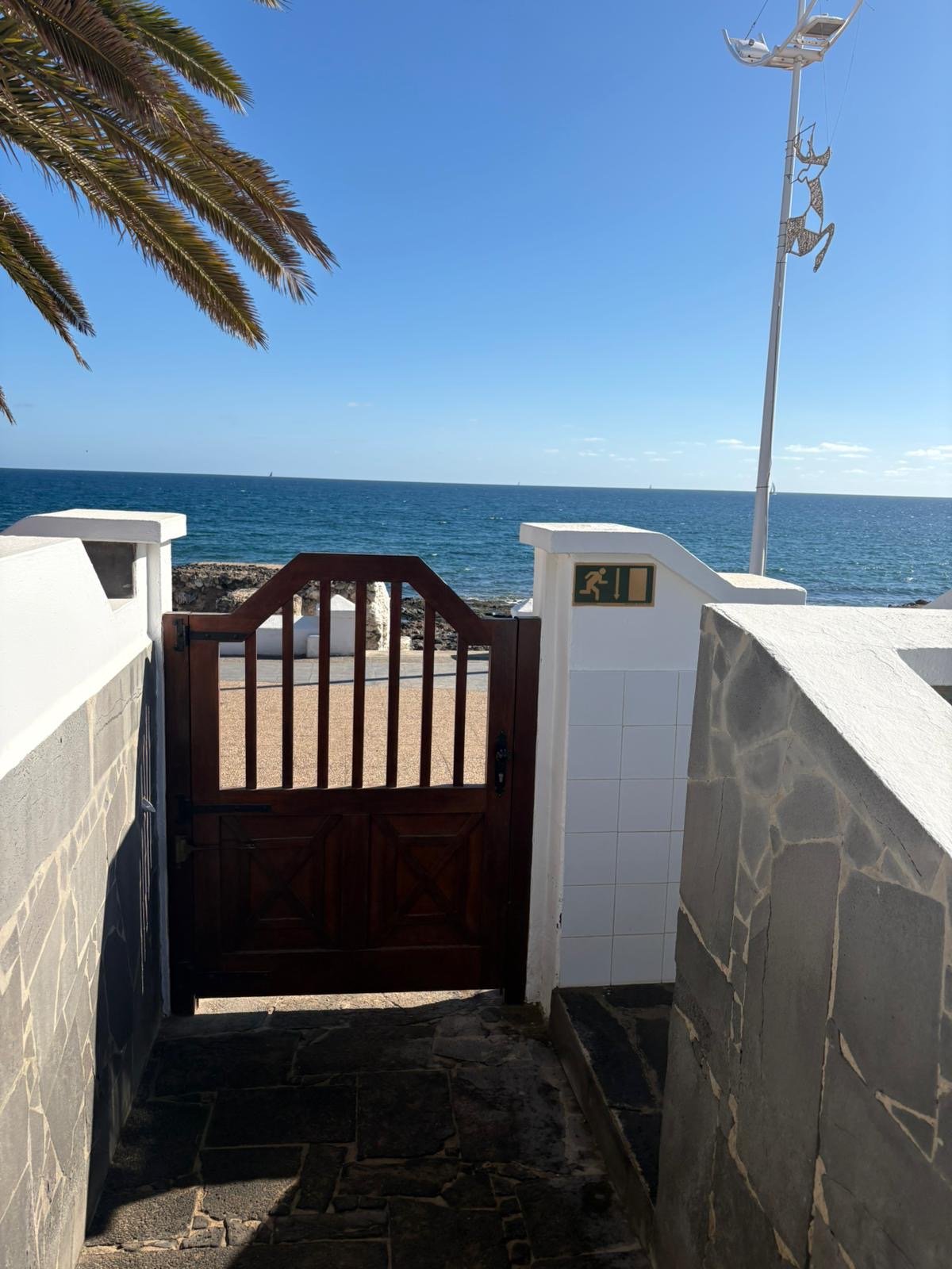 Wooden gate leading to ocean view with palm tree, stone walls, and a clear blue sky.