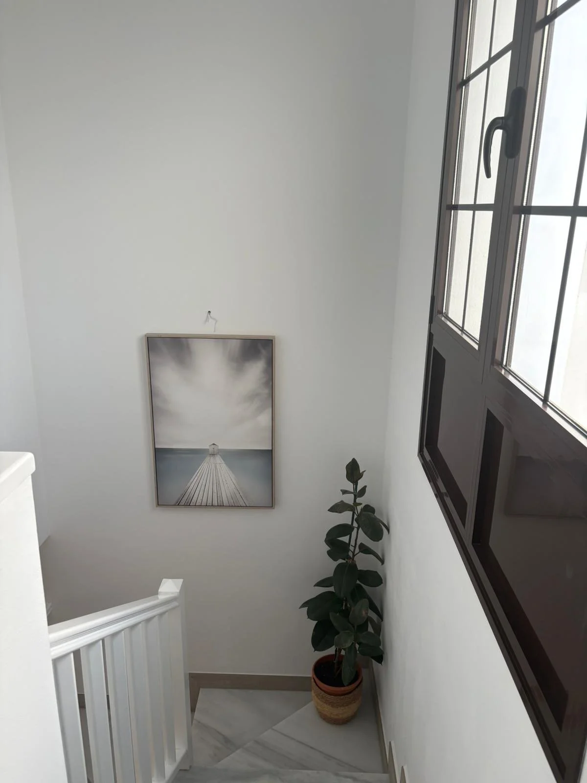 Staircase with white walls, a framed photo of a pier leading to water, a potted plant, and a window with dark frame.