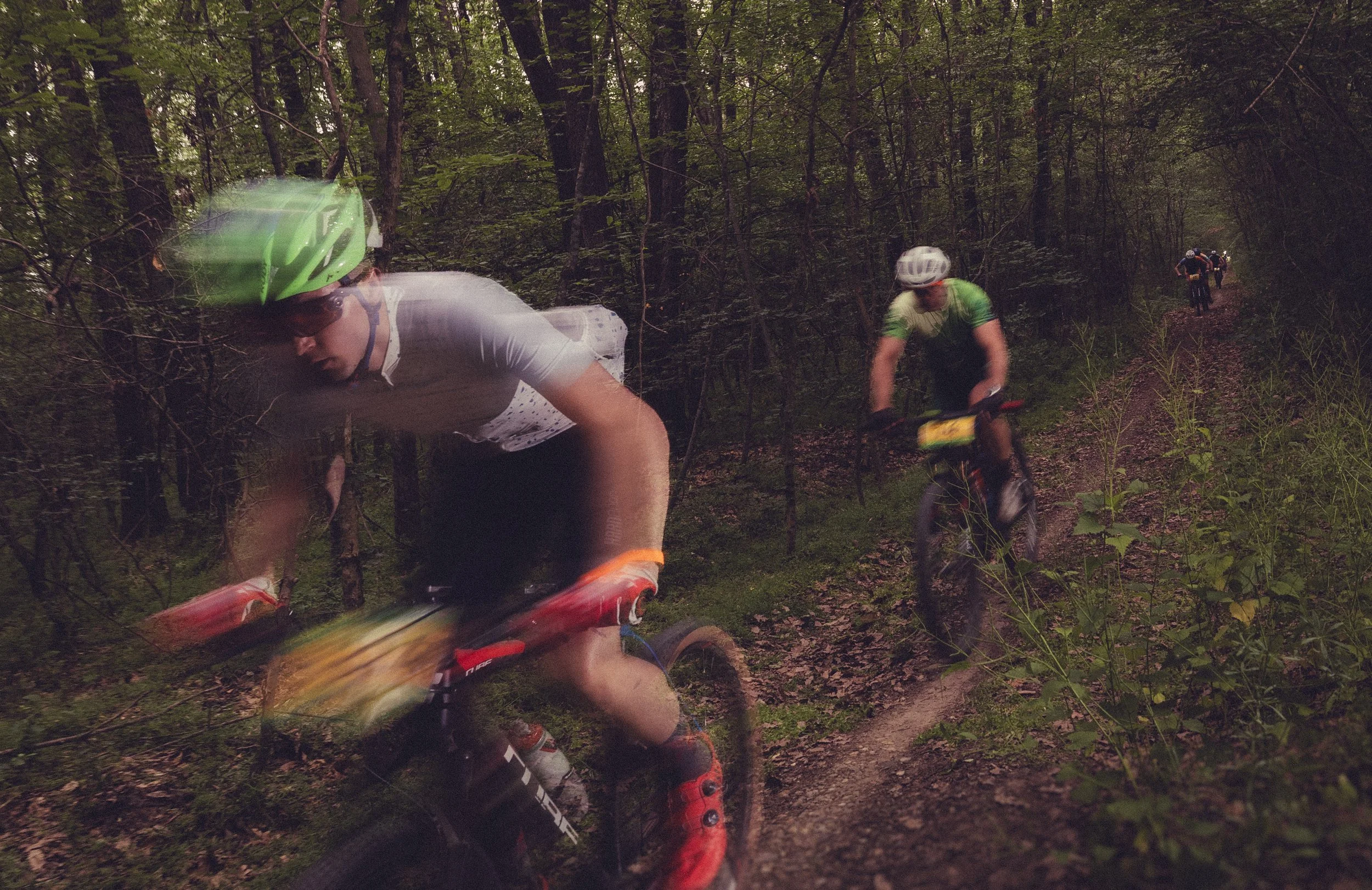 Group of mountain bikers riding through a wooded trail, with one rider in the foreground wearing a green helmet and white shirt, and others behind on a narrow dirt path surrounded by trees and foliage.