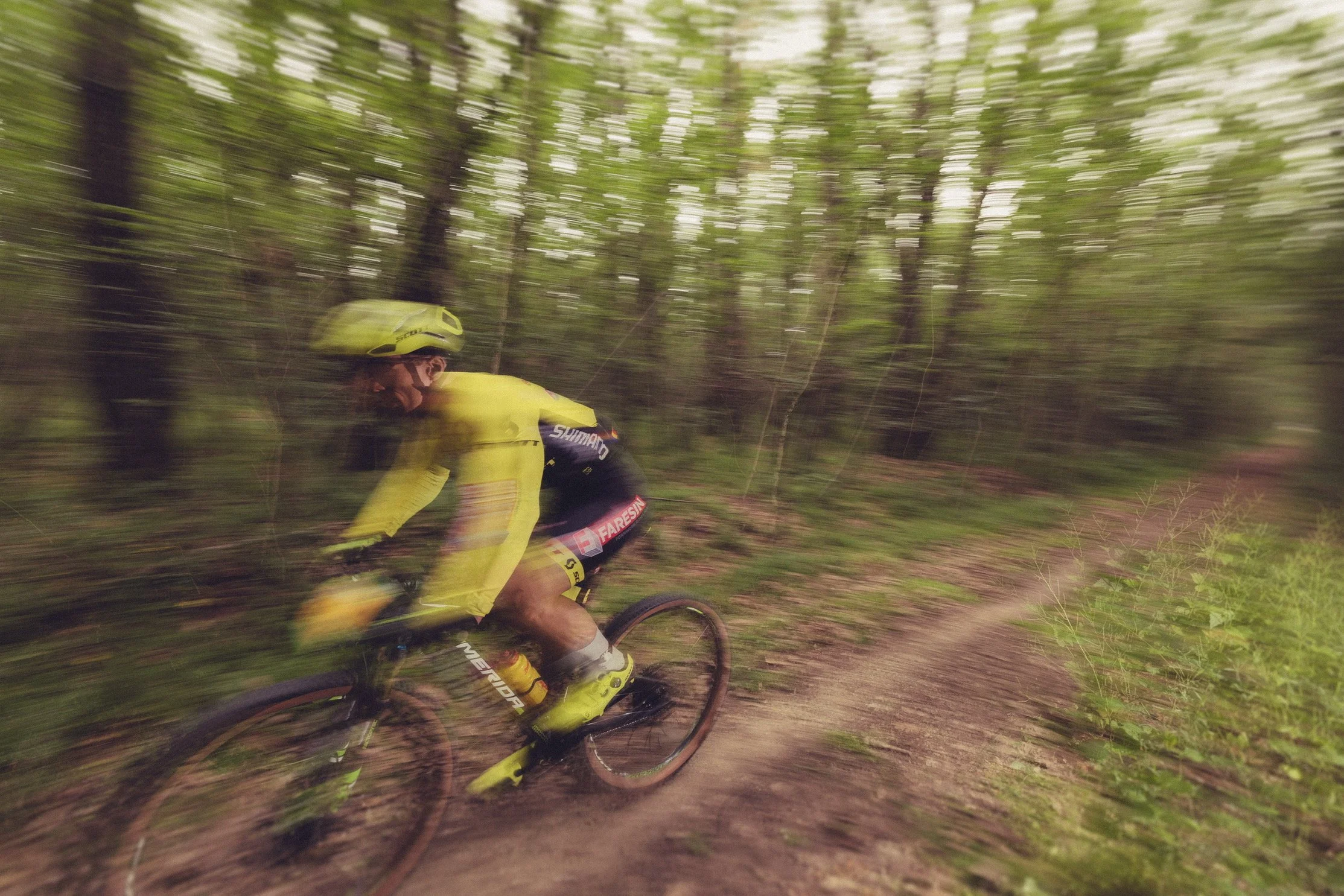 A mountain biker wearing a yellow helmet and yellow gear riding on a dirt trail through a green forest. The photo has a motion blur effect.