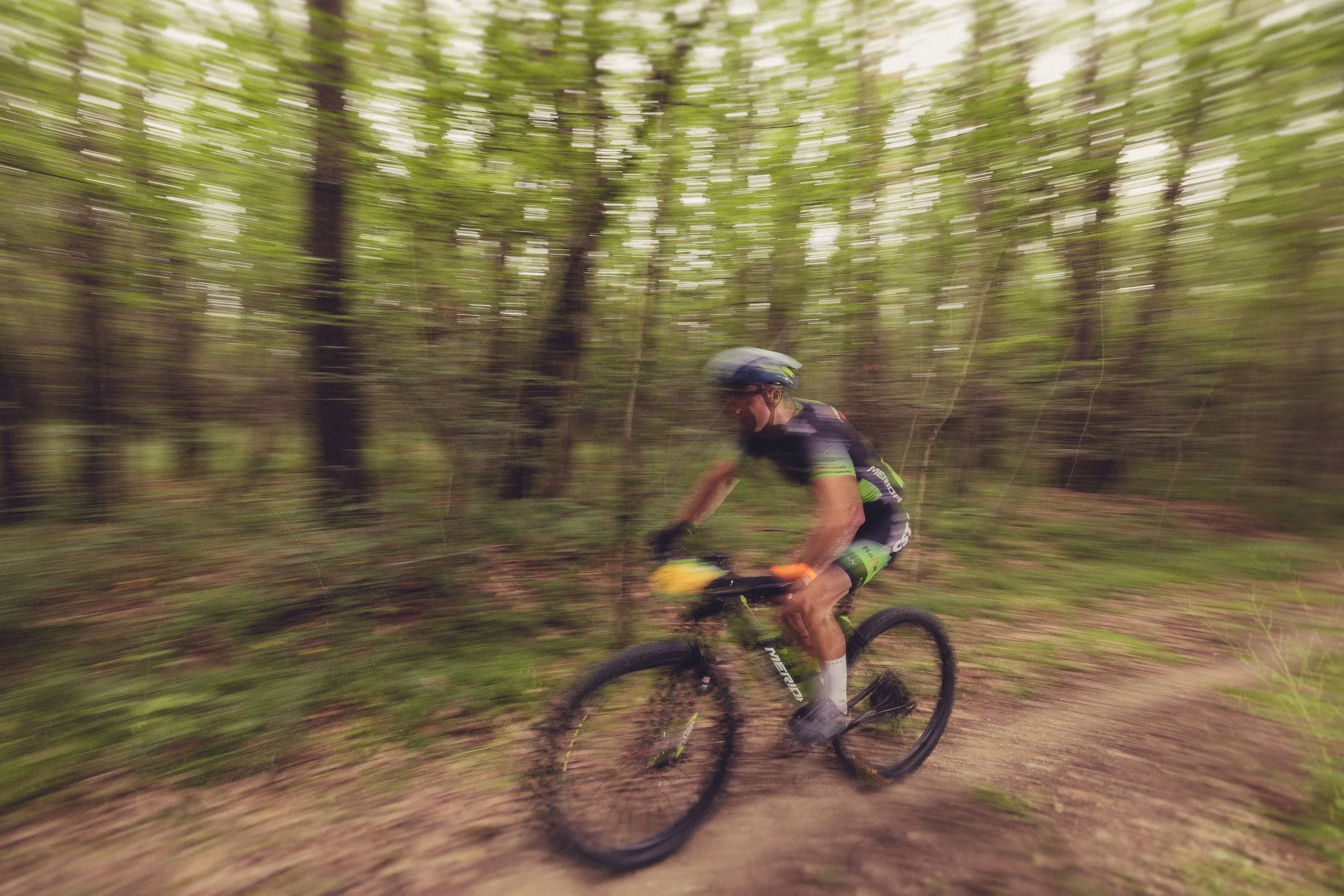 A person mountain biking on a trail through a wooded forest, with motion blur indicating fast movement.