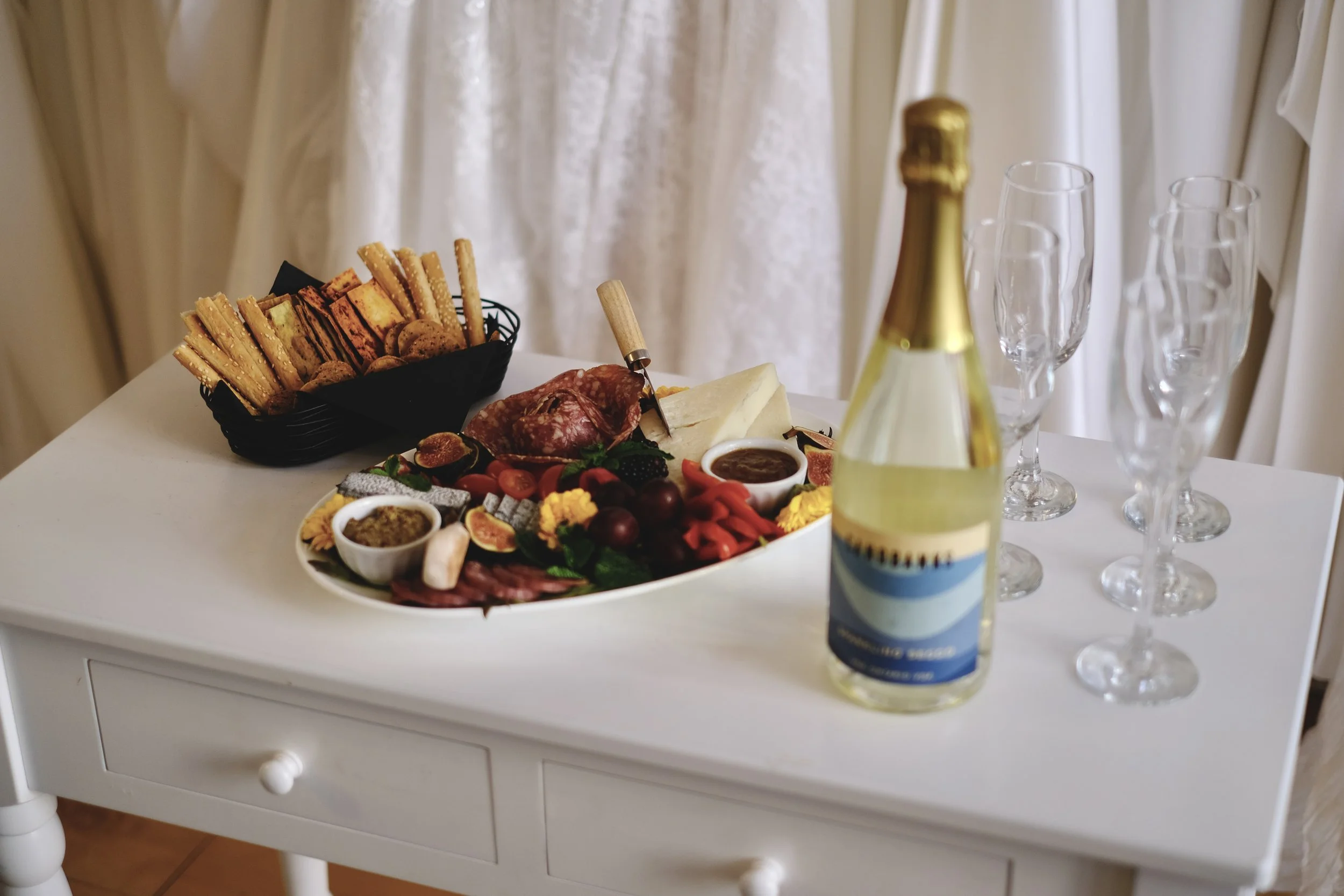 Charcuterie board with cheese, crackers, strawberries, and snack foods on a lace tablecloth, with a table of assorted dishes and desserts in the background.