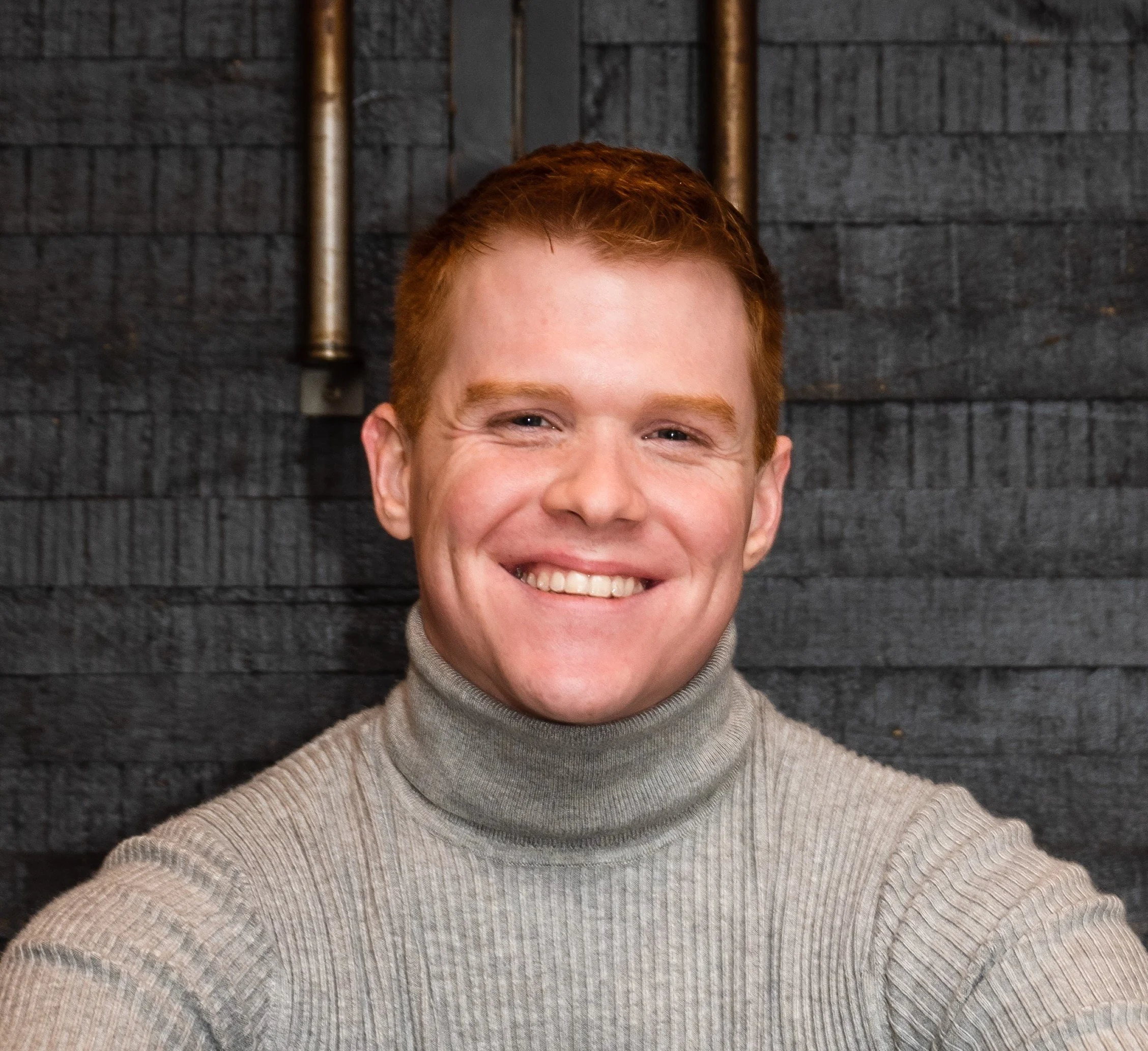 A young man with red hair and a big smile wearing a gray turtleneck sweater, standing in front of a dark gray brick wall.