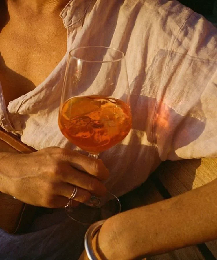Close-up of a hand holding an orange spritz cocktail outdoors at sunset
