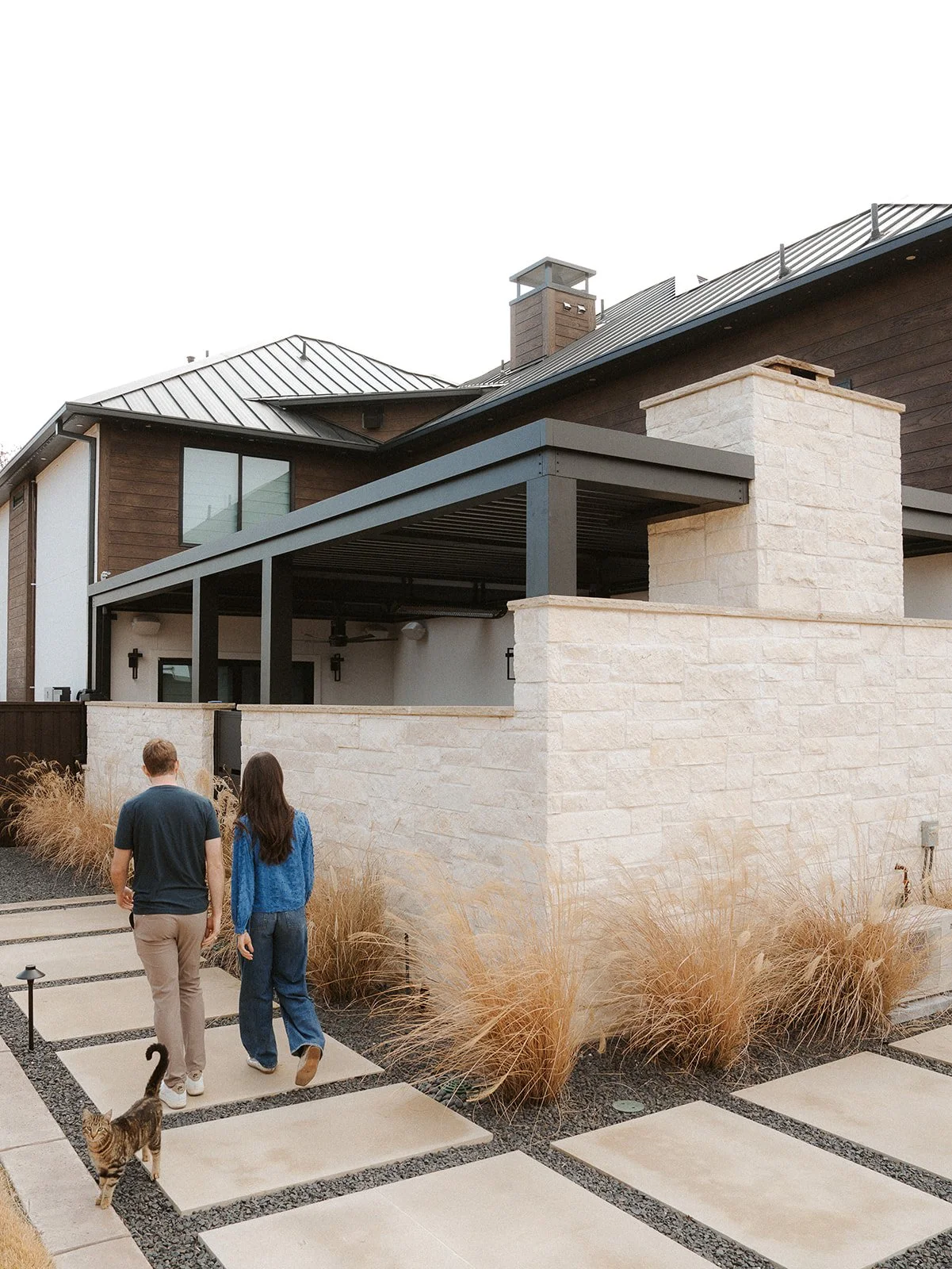 A man installing a black metal railing on a stone patio in front of a modern home under cloudy skies, with construction materials and tools nearby.