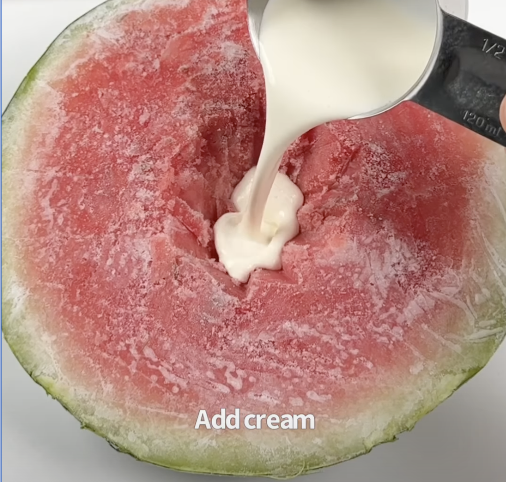 Cream being poured from a measuring cup into a small well carved in the center of a frozen watermelon half.