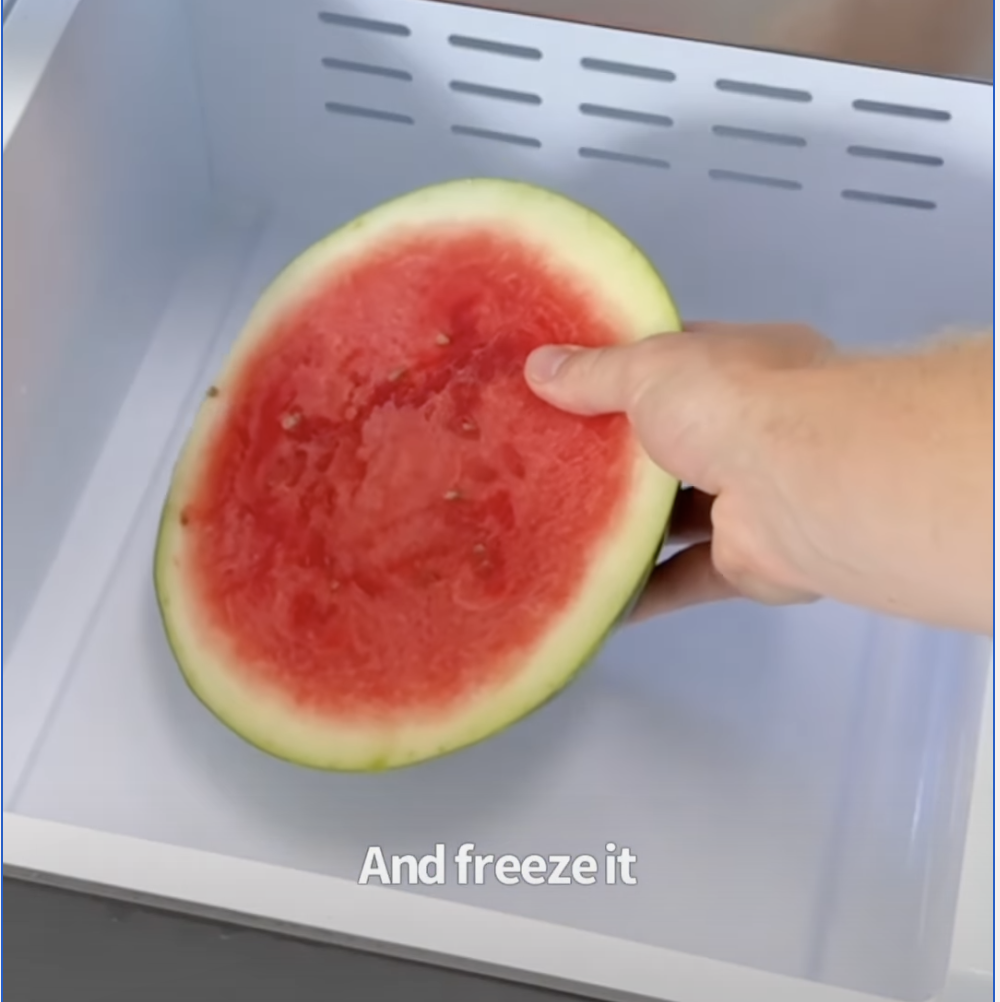 Hand placing a halved watermelon cut-side up into a freezer drawer.
