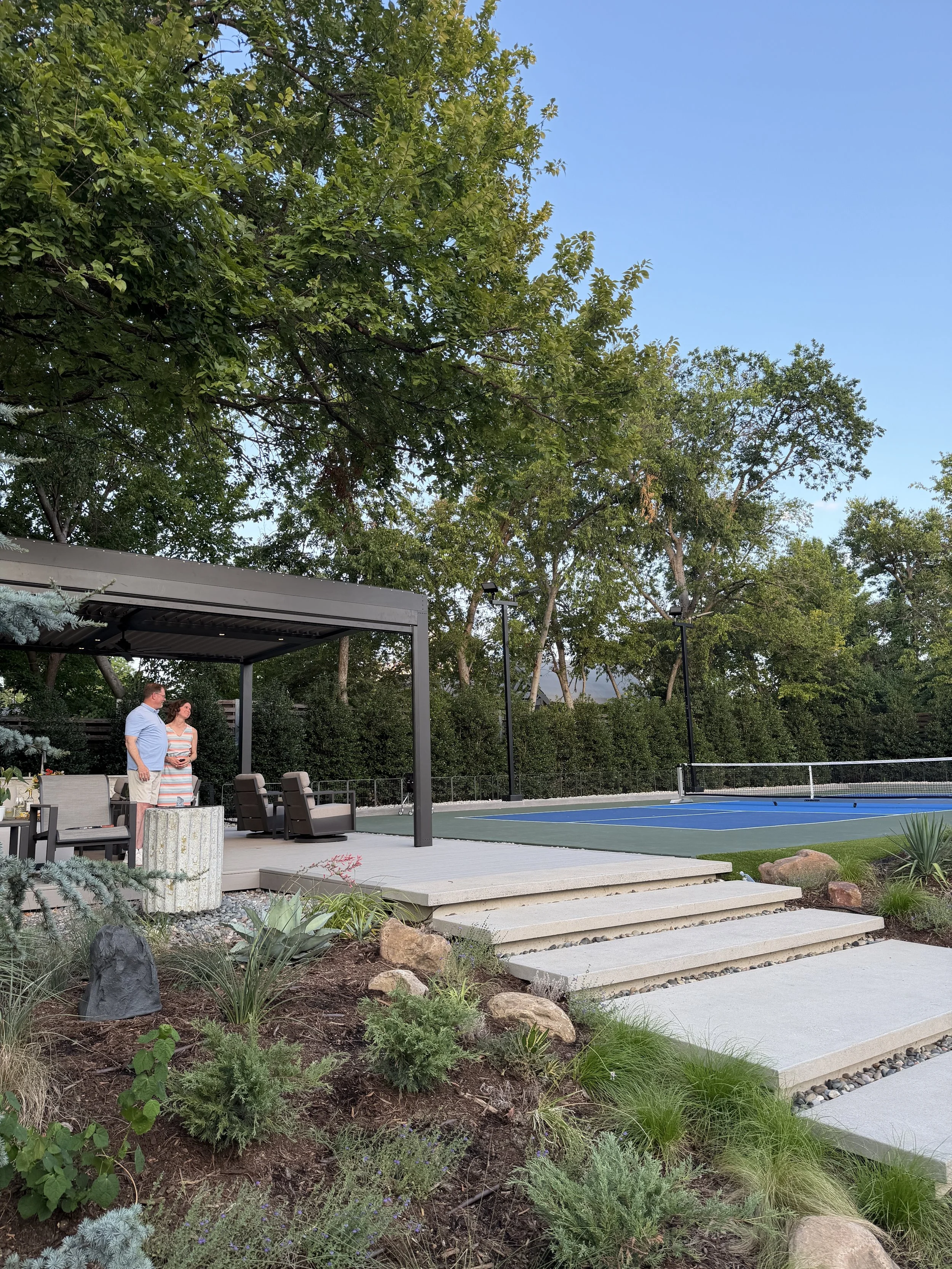 Couple standing under a black StruXure motorized pergola on a stone deck, with steps leading down to a custom backyard pickleball court framed by mature trees and native landscaping.