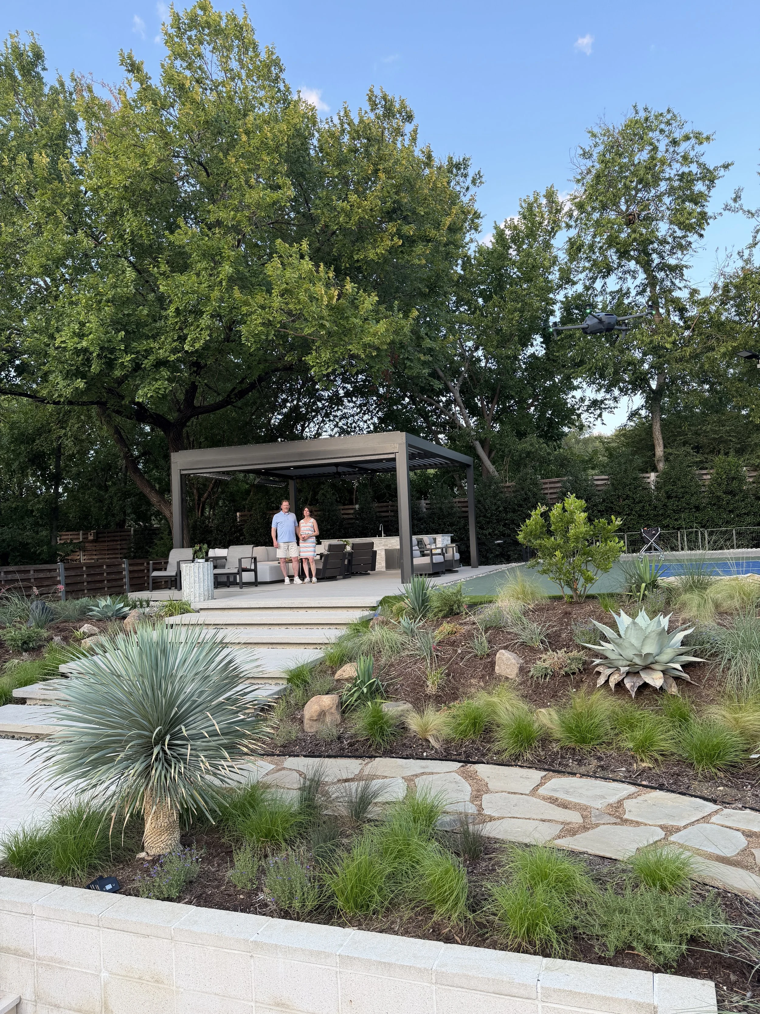 Couple standing under a finished outdoor pergola overlooking a landscaped backyard and pool
