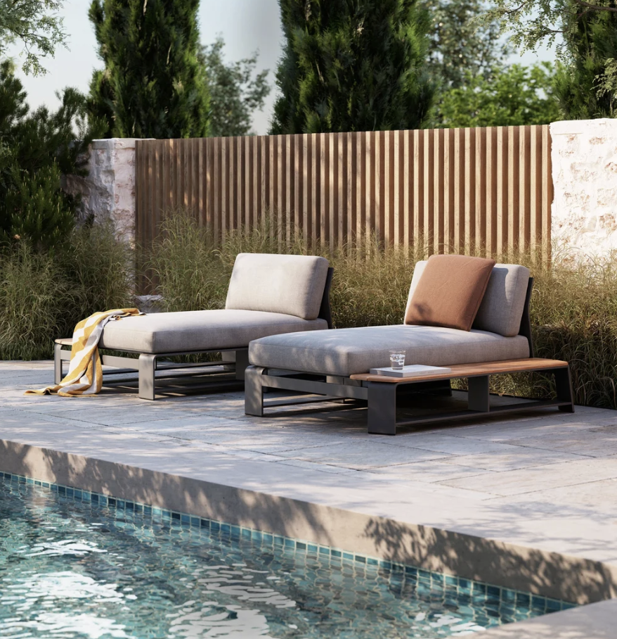 Two low-profile Rose Casual chaise loungers in taupe fabric with a rust accent pillow and striped throw blanket, arranged poolside on a stone deck beside a teak side table with a glass and a book.