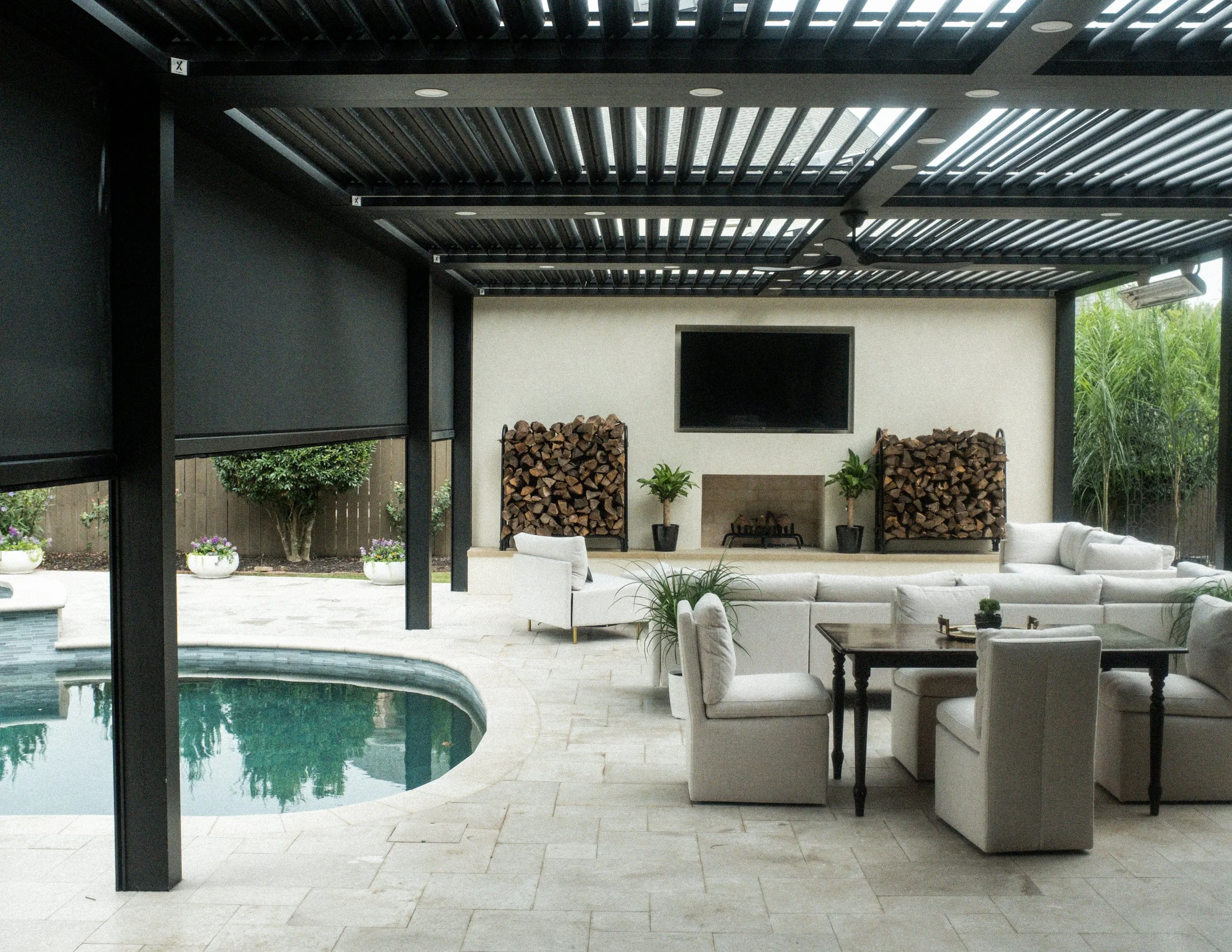 Fully built outdoor living room under a black StruXure motorized louvered pergola, featuring a built-in fireplace with mounted TV, firewood storage, a white sectional sofa and lounge chair