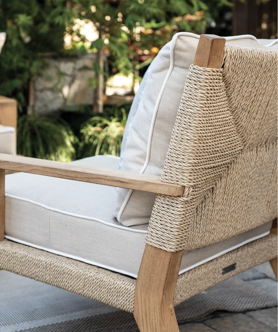 Close-up of a Rose Casual lounge chair's hand-woven rope back and teak wood frame, with cream cushions and white piping, garden backdrop softly blurred.