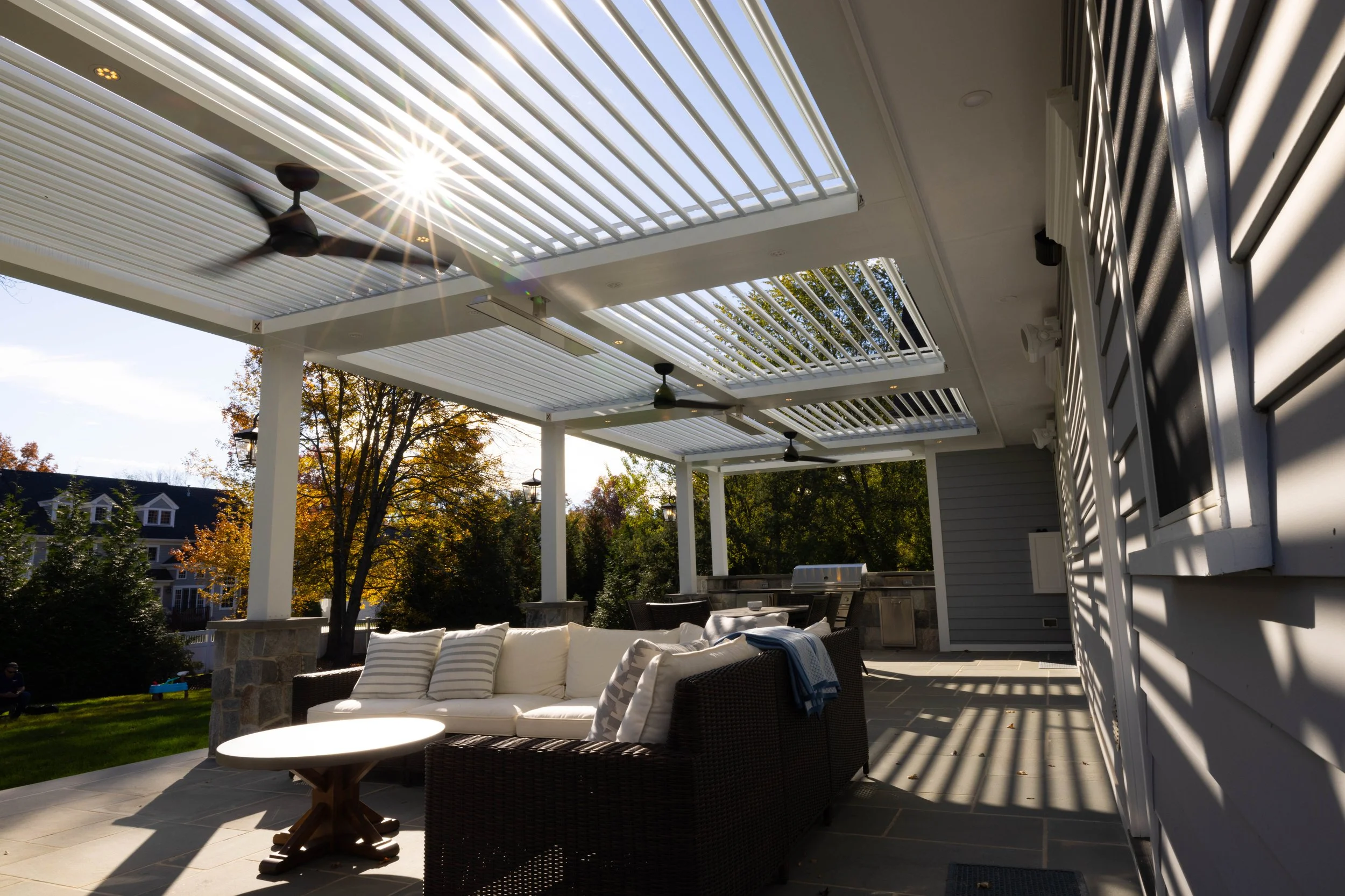 Covered patio with white ceiling slats, ceiling fans, outdoor furniture including a sofa with cushions, a small table, a barbecue grill, and surrounding trees with fall foliage.