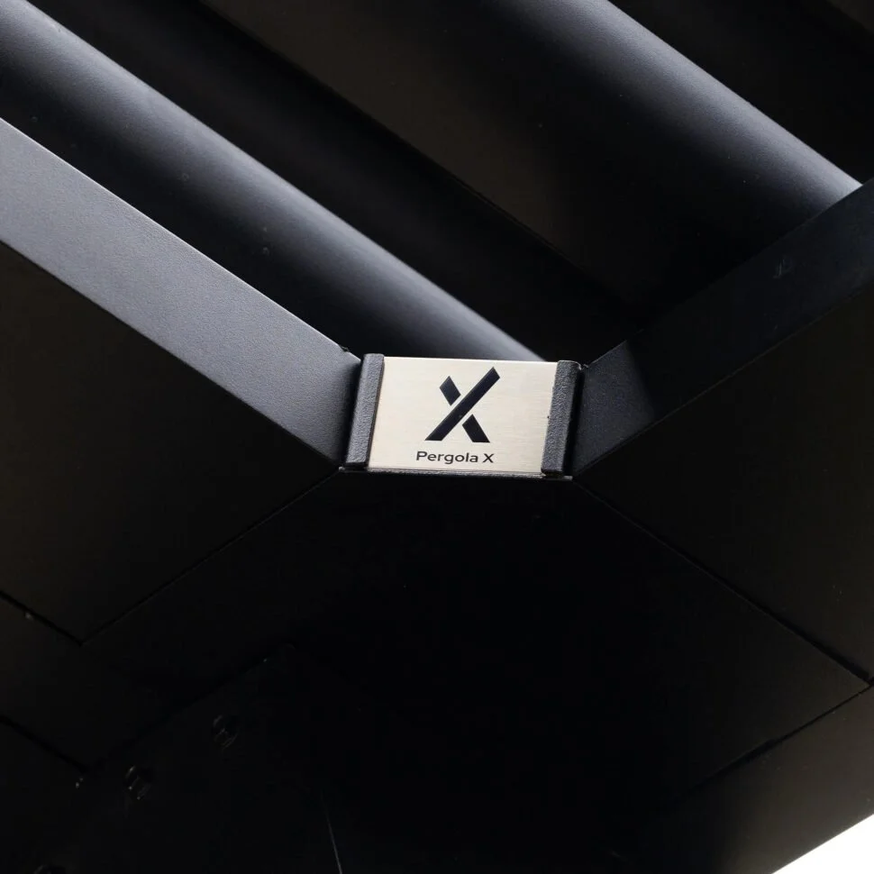 Close-up of a black outdoor pergola with a silver metal label reading 'Pergola X' and a stylized logo in black.