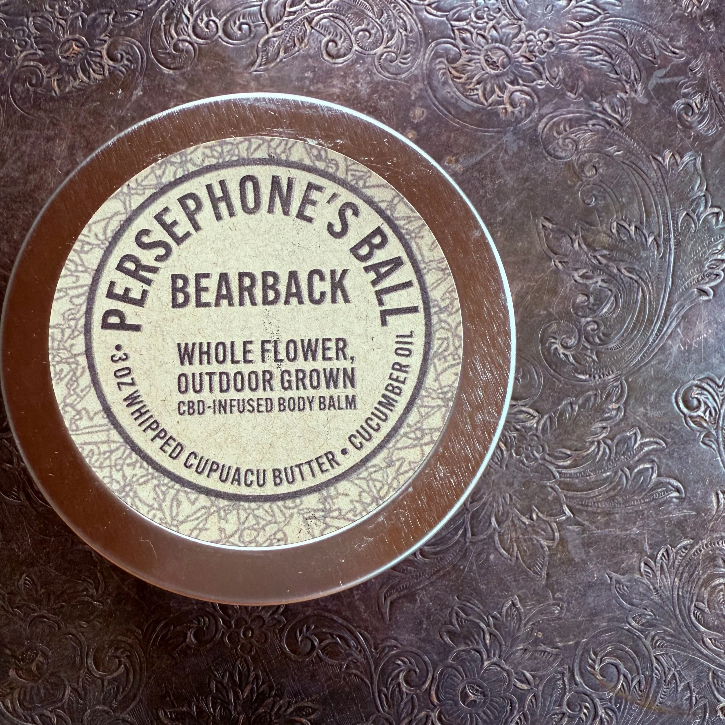 Circular label on a dark metallic surface, reads 'PERSEPHONE'S BAIL BEARBACK WHOLE FLOWER, OUTDOOR GROWN CBD-INFUSED BODY BALM, WHIPPED CUPUACU BUTTER, CUCUMBER OIL'.