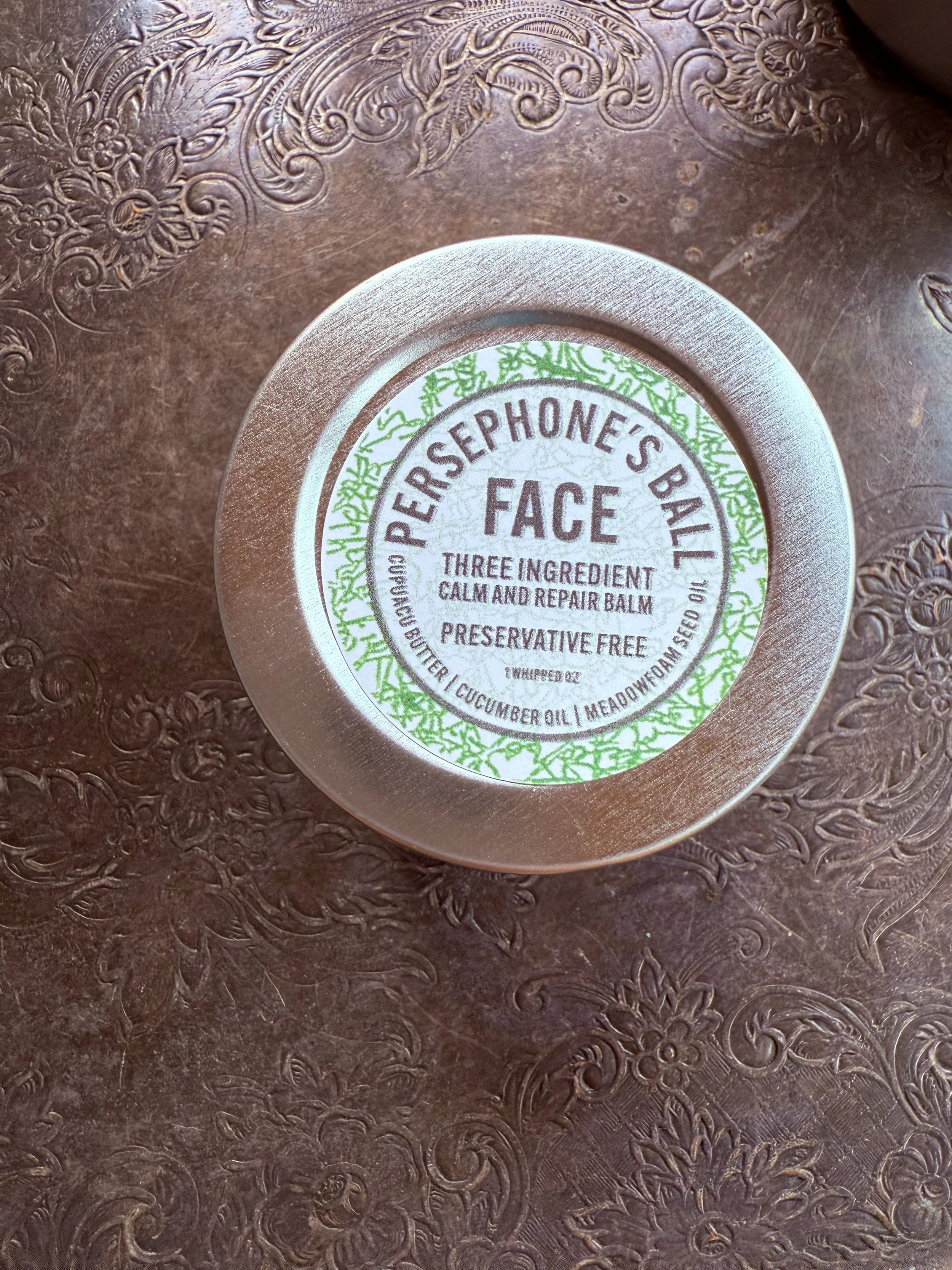 A round tin container labeled 'Sephone's Balm Face' resting on an ornate, embossed copper surface. The label mentions it is a calming and repairing balm made with butter, cucumber oil, and meadowfoam seed oil.