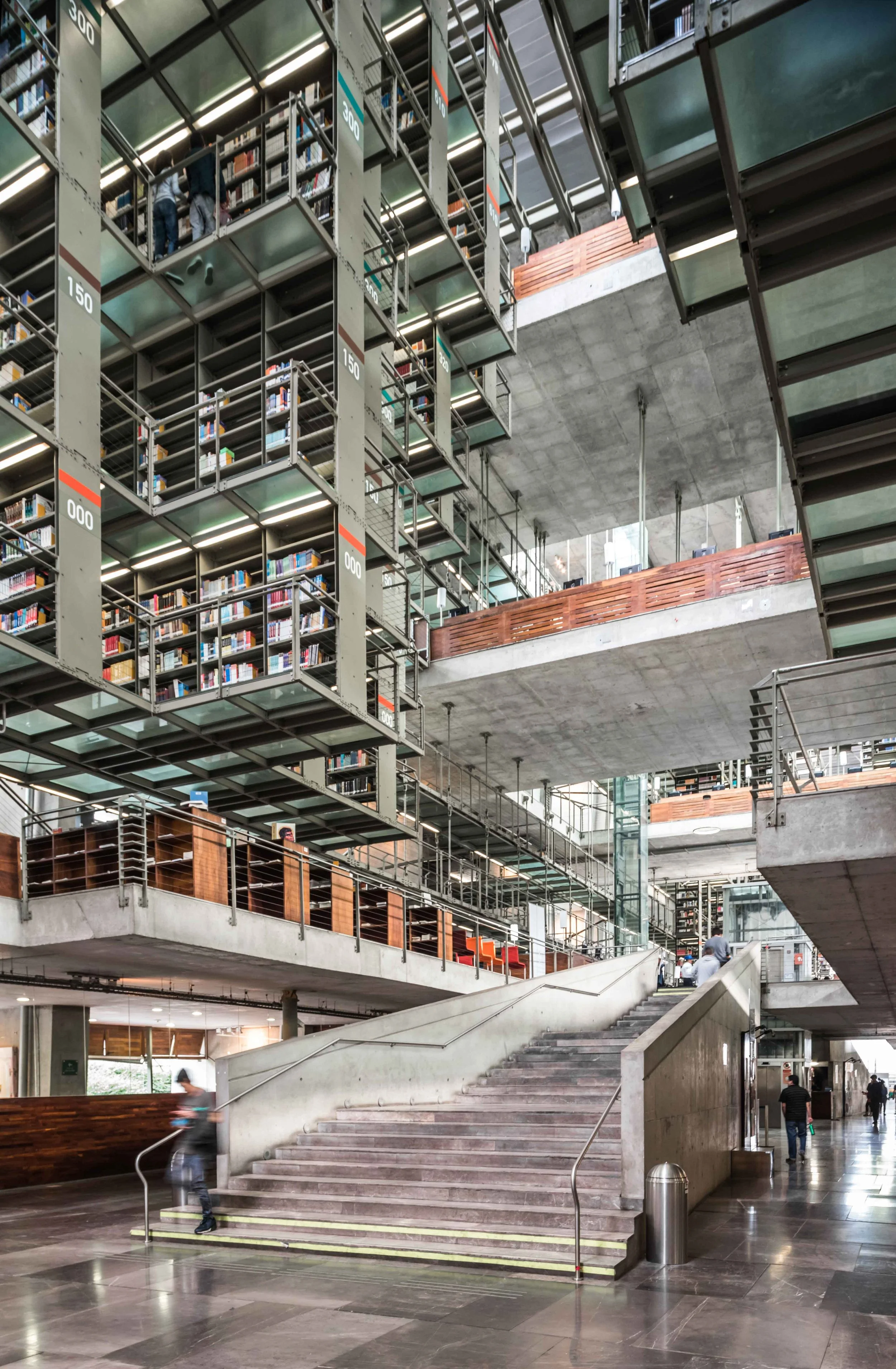 Interior moderno de una biblioteca con escaleras, estanterías con libros, áreas abiertas y estructuras de concreto y metal.
