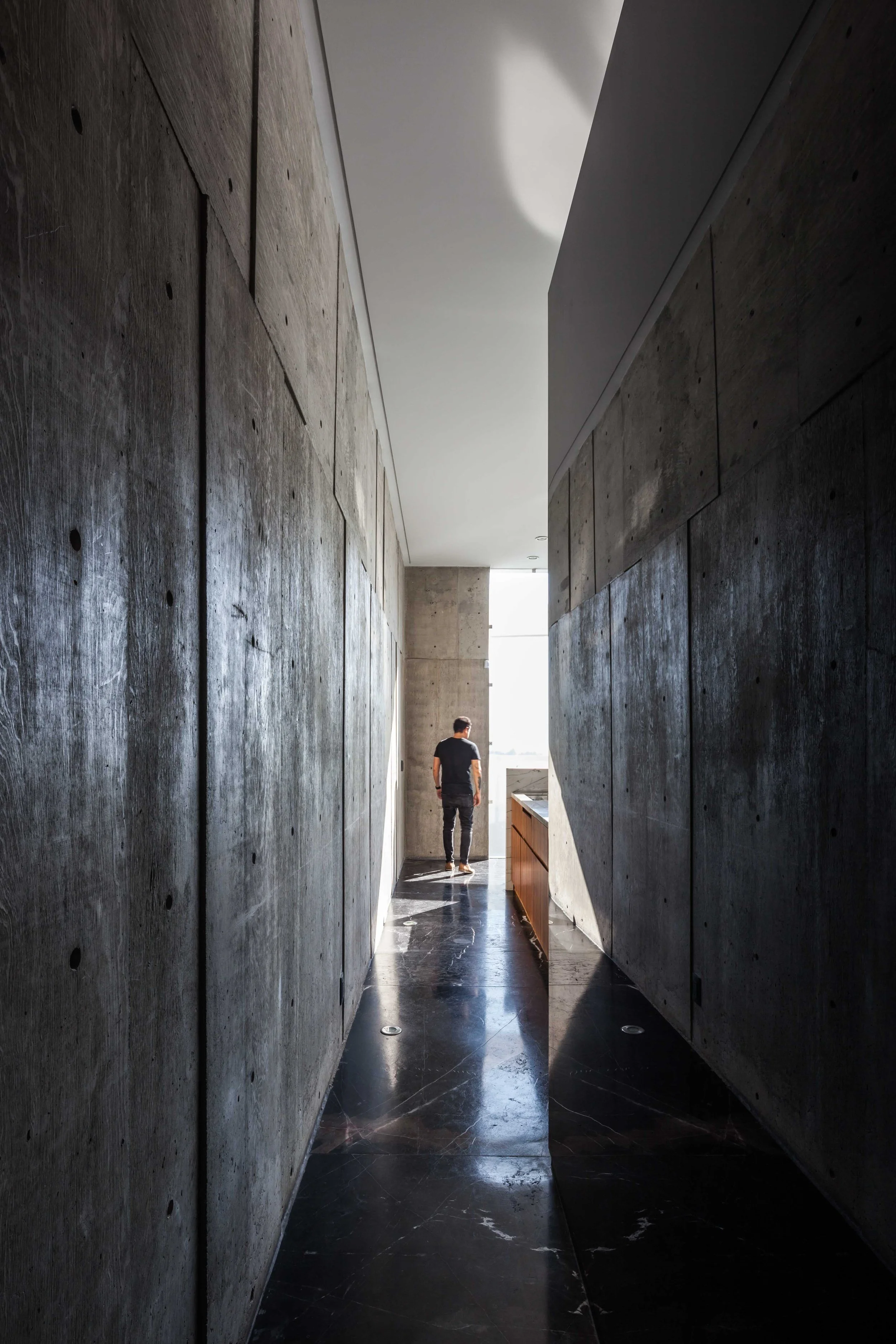 Pasillo estrecho con paredes de concreto, suelo de mármol negro y una persona de espaldas mirando hacia una ventana en el fondo.