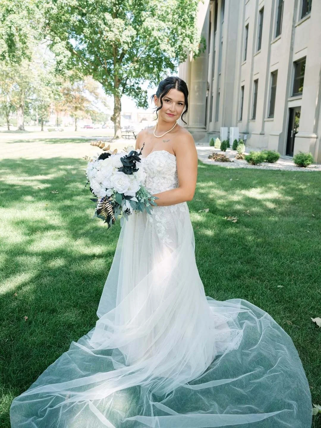 Bride standing on a green lawn with a floral bouquet and wedding dress blowing in the wind