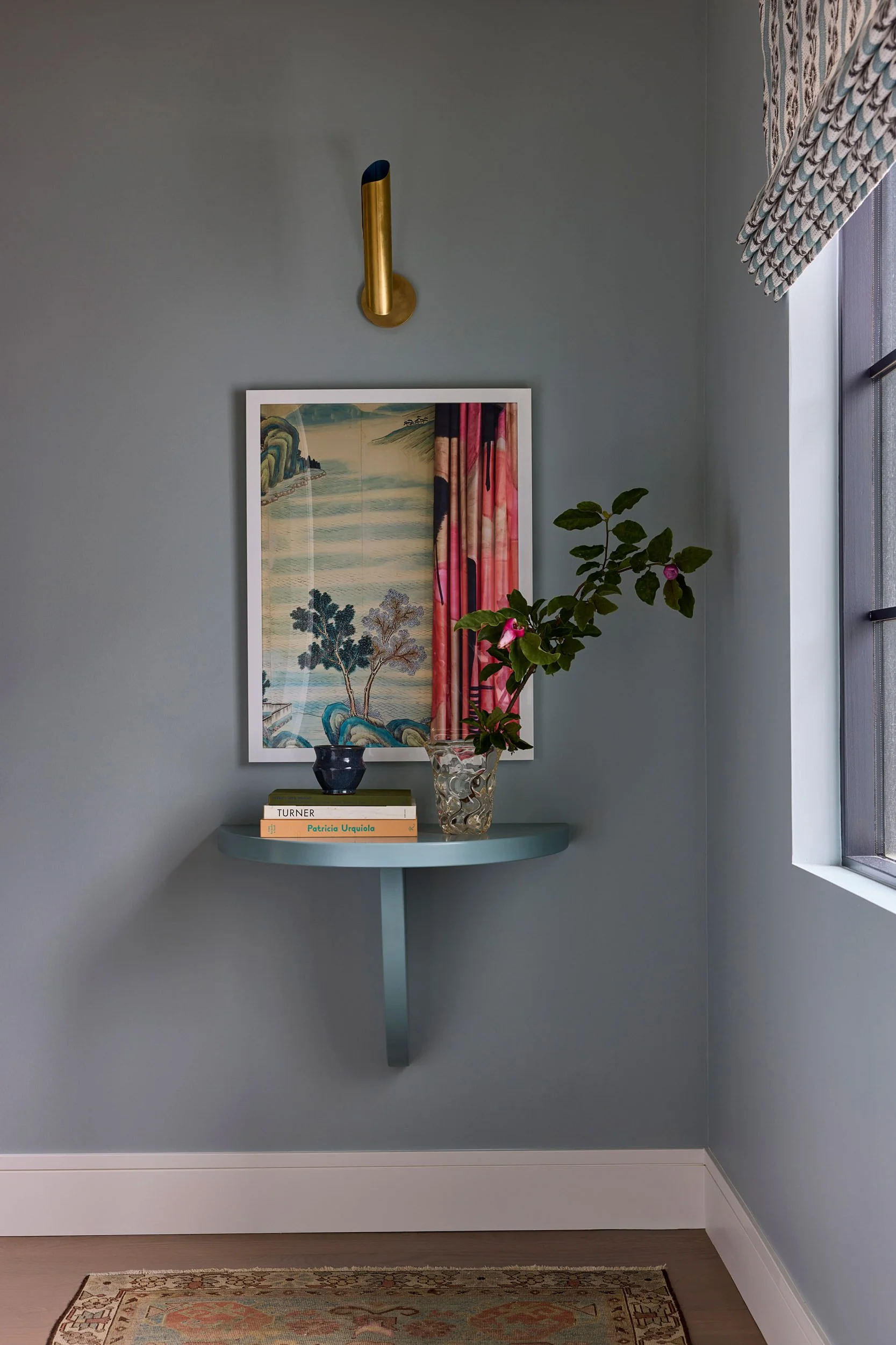 Framed colorful artwork above a light blue demilune table with decorative books and flowers, set against soft blue-gray walls with a brass wall sconce and patterned window shade.