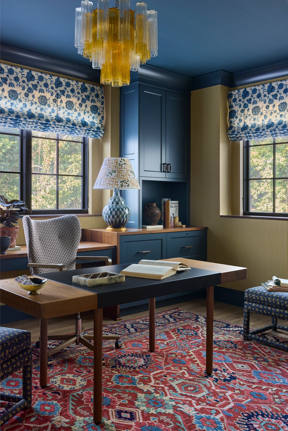 Home office featuring wood desk, blue cabinetry, blue floral Roman shades, an amber glass chandelier, and a colorful red patterned rug.