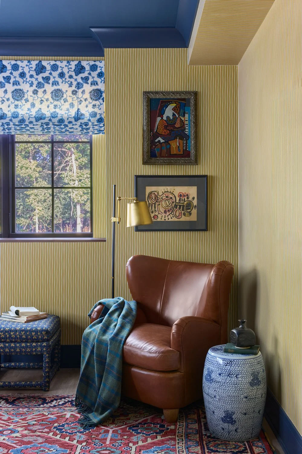 Home office reading nook featuring a leather armchair, striped yellow wallpaper, blue floral Roman shade, framed artwork, and a colorful red patterned rug.