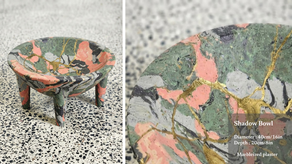 A small, colorful marbleized shadow bowl with three legs, featuring a pink, green, black, and gray pattern with gold veins, placed on a speckled surface.