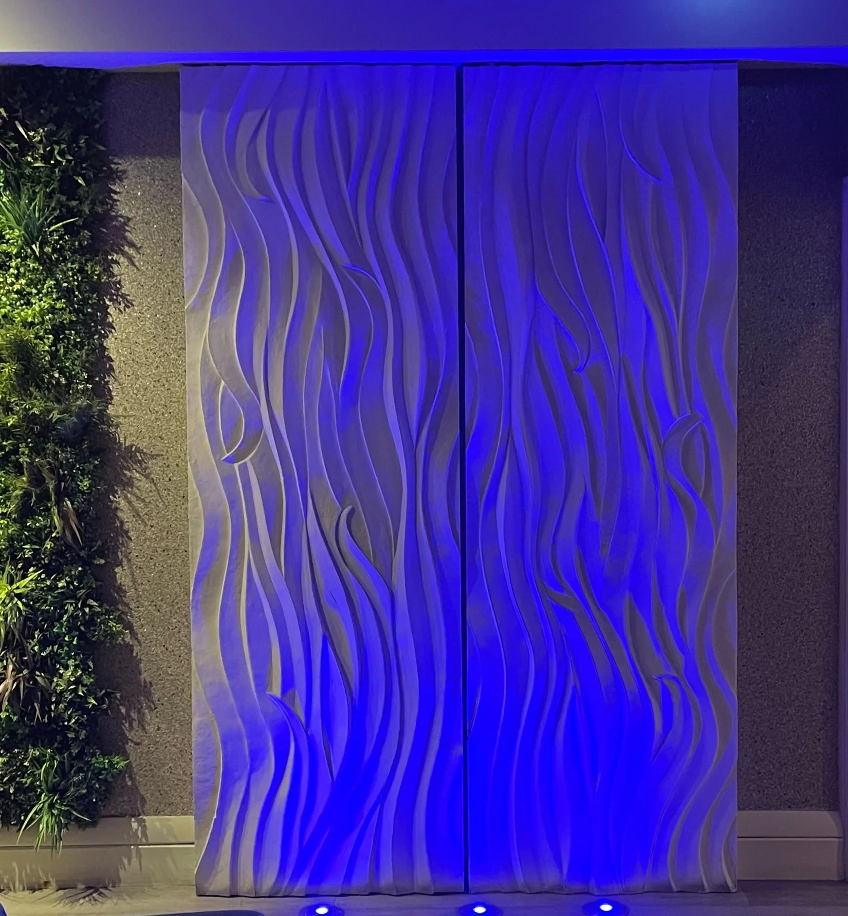 Smooth Sand Ripple wall panel lit in deep blue in a hotel corridor beside planting