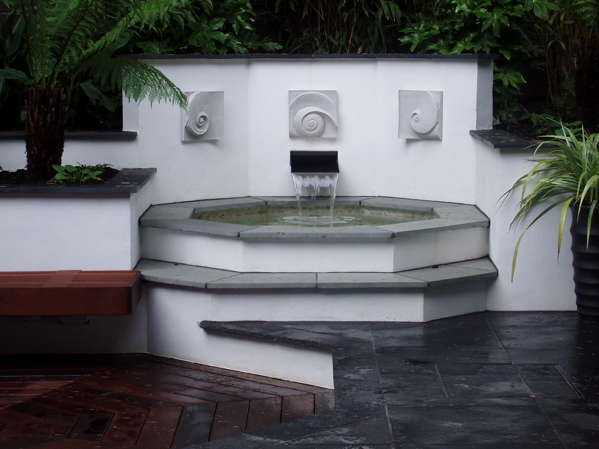 Moonphases Three wall sculpture above a white water feature spilling into a pool in a planted courtyard