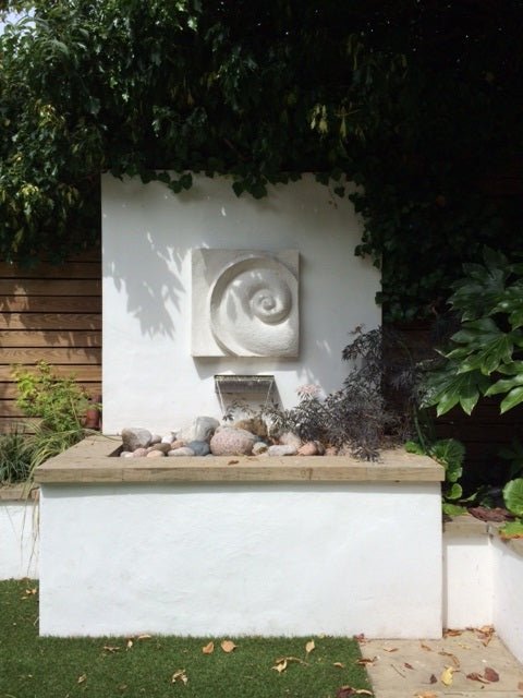 Spiralmoon wall sculpture mounted on a white garden wall above a pebble water feature and climbing greenery