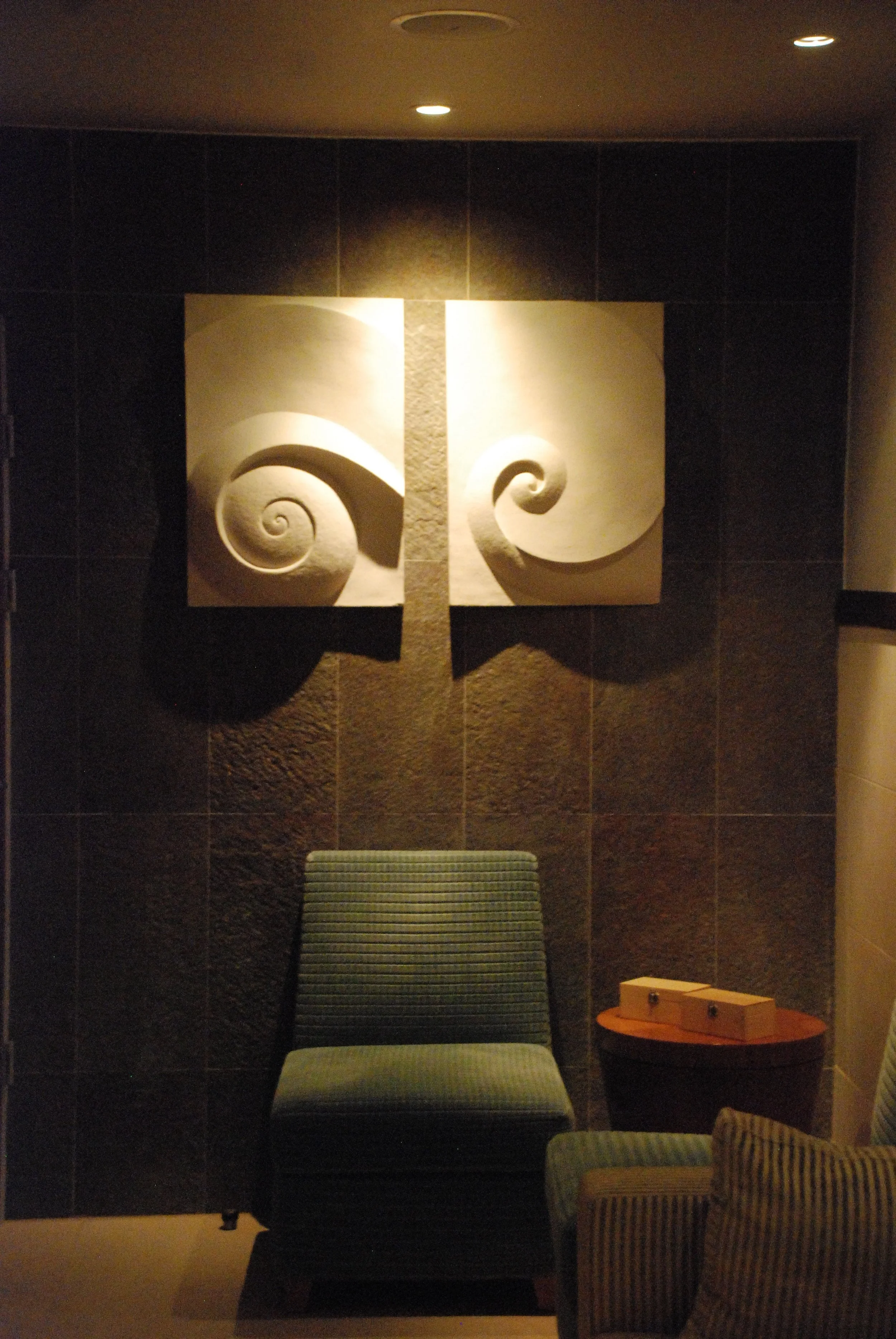 Two Moonspiral wall sculpture panels lit on the wall of a dark interior above a low armchair